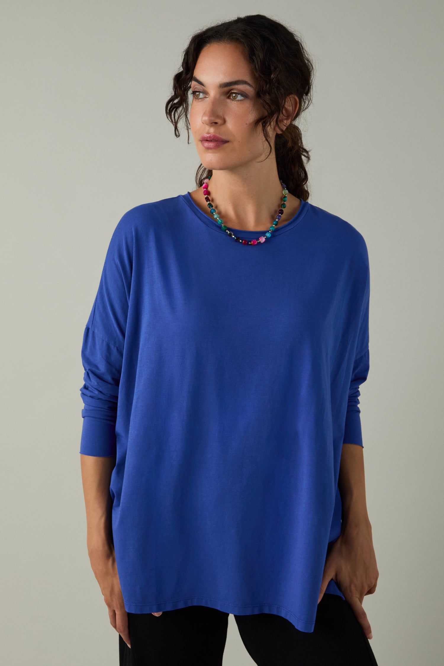 A woman wears the Cotton Jersey Relaxed Top with a comfortable fit, paired with black pants and a beaded necklace, standing against a neutral background. This top is a true wardrobe essential for effortless style.