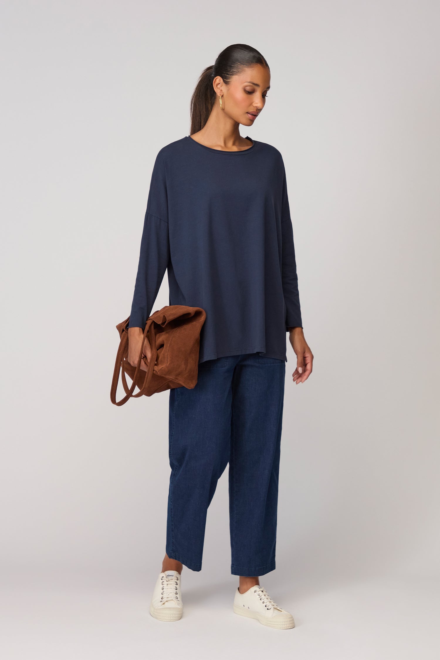 A woman stands against a plain background wearing the Cotton Jersey Relaxed Top in navy, loose blue jeans, white sneakers, and holding a brown bag—an effortless wardrobe essential.