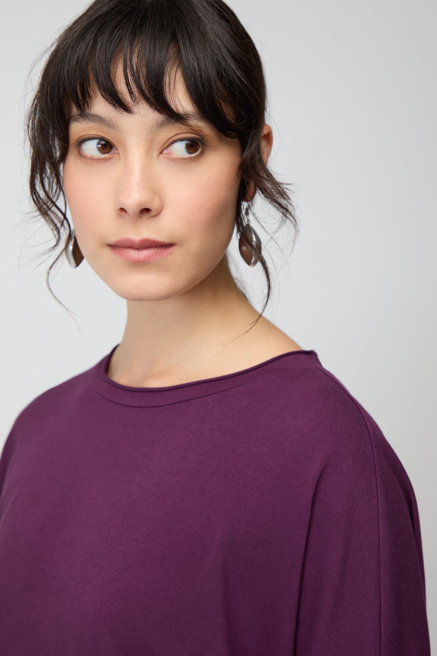 Woman with wavy dark hair and bangs wears the Cotton Jersey Relaxed Top in purple, looking left against a plain light background—an effortlessly comfortable wardrobe essential.