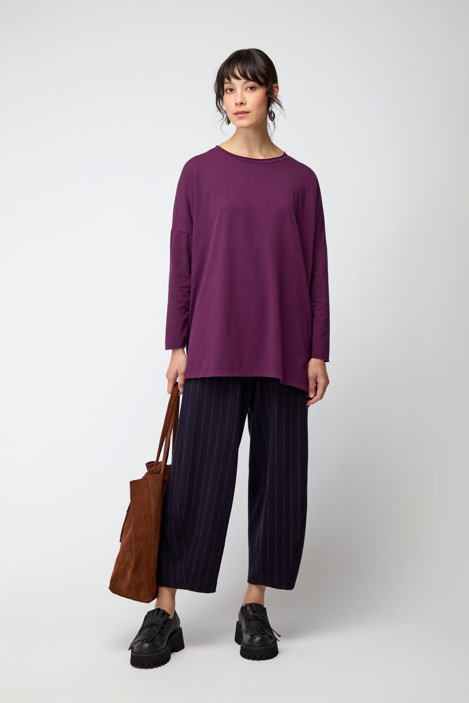 Woman stands against a plain background wearing the Cotton Jersey Relaxed Top in purple, wide-leg striped dark pants, black shoes, and holding a brown tote bag—an effortless essential offering relaxed comfort.