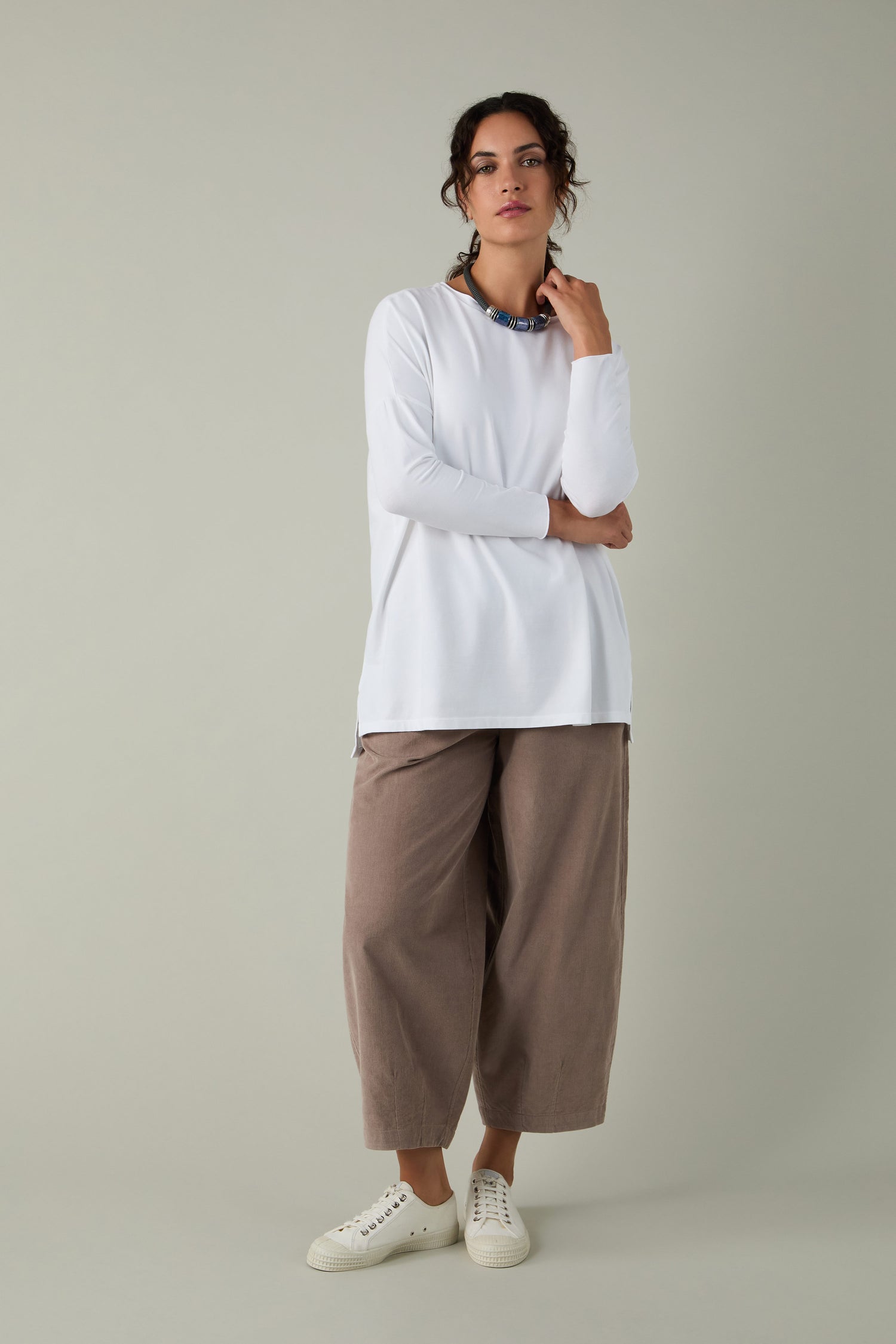 A person stands against a plain background wearing the Cotton Jersey Relaxed Top, styled with loose brown pants, white sneakers, and a colorful necklace—a relaxed essential for any wardrobe.