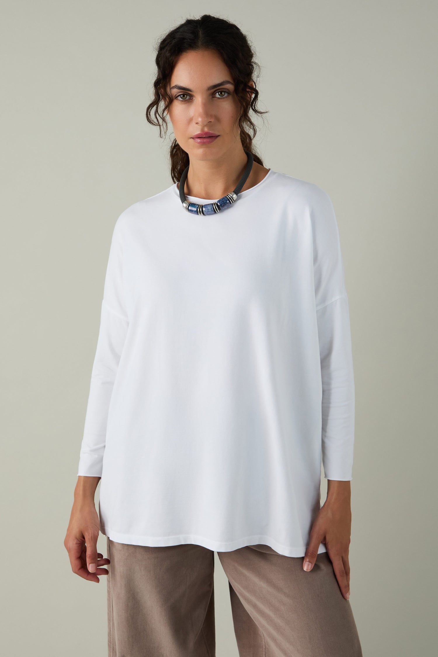 A woman stands against a plain light background wearing the Cotton Jersey Relaxed Top, styled for comfort, paired with beige pants and a chunky necklace.