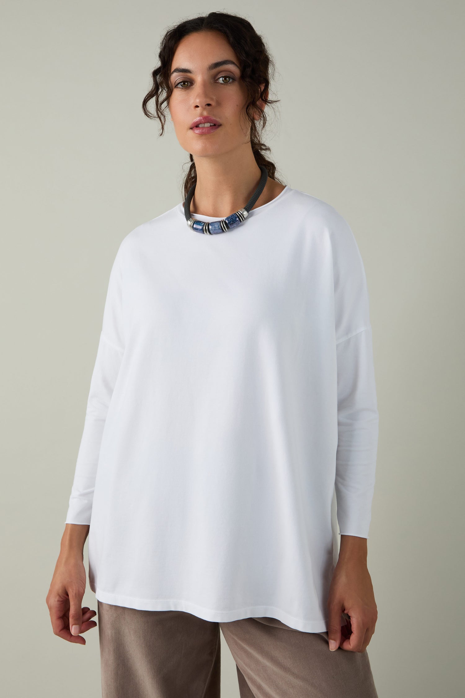 A woman faces the camera in a Cotton Jersey Relaxed Top, styled with taupe pants and a chunky blue-silver necklace against a plain background—a versatile wardrobe essential.