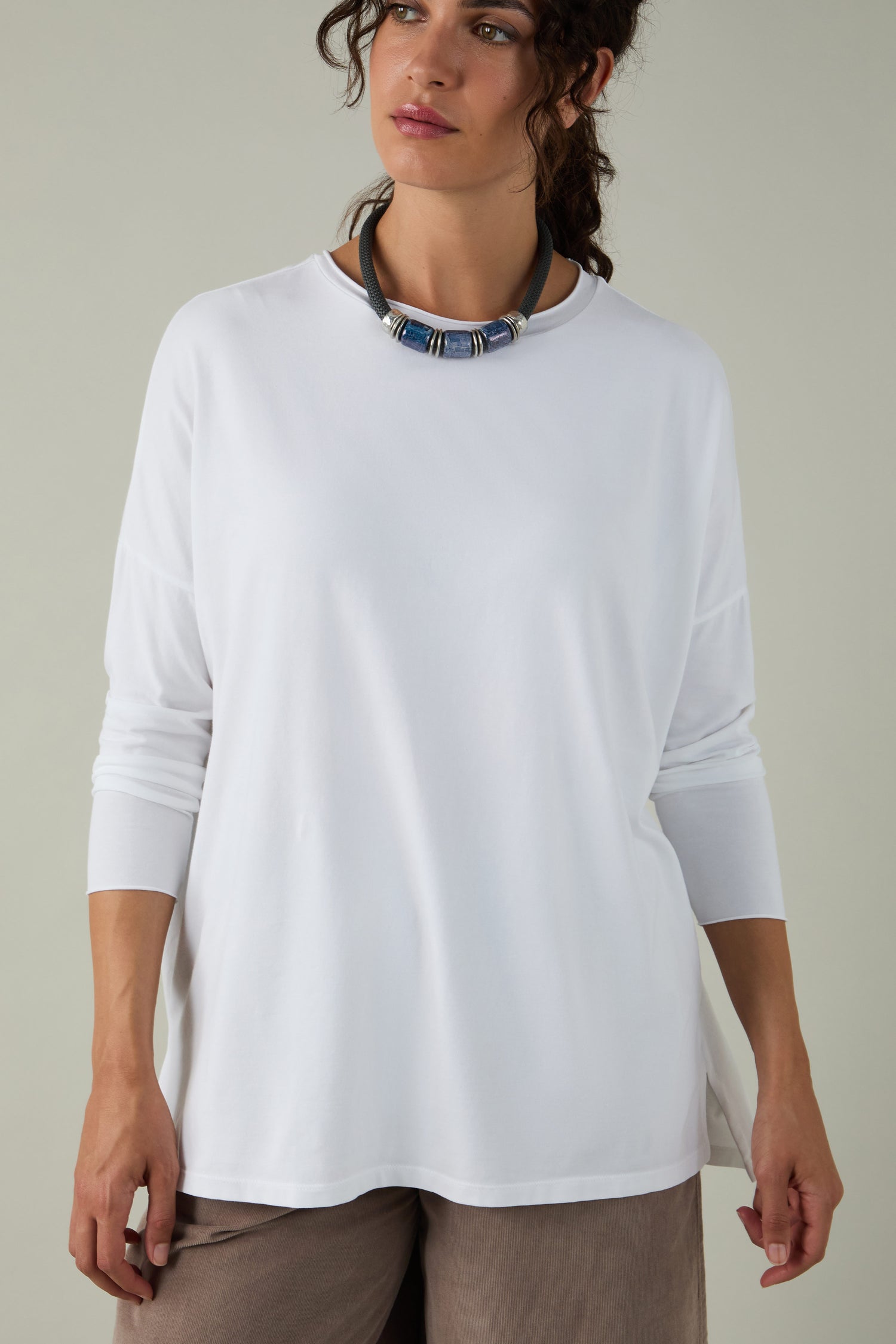 A woman stands against a plain background wearing the Cotton Jersey Relaxed Top, styled with beige pants and a chunky necklace. This comfortable-fit top is a true wardrobe essential.