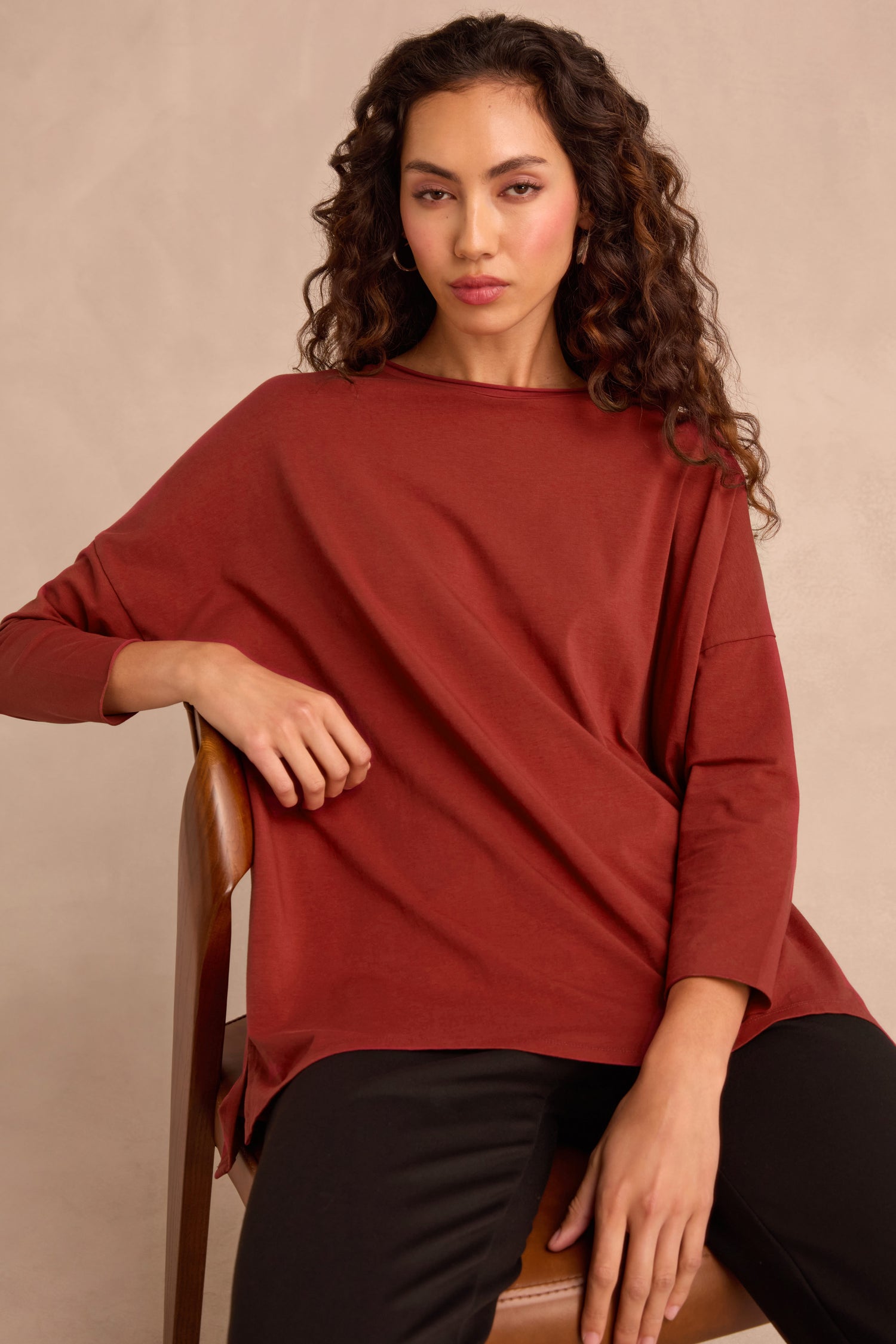 A woman with long, curly hair sits on a wooden chair, wearing the Conker Cotton Jersey Relaxed Top in rust and black pants against a neutral background.