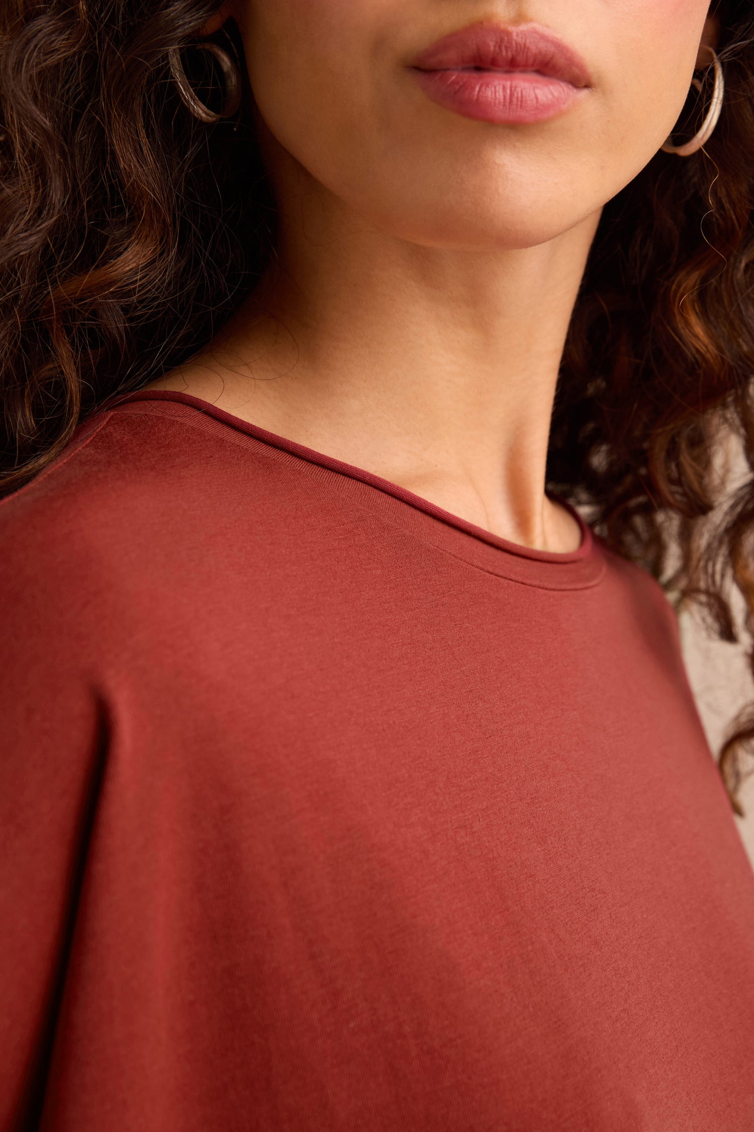 A woman with curly brown hair and hoop earrings wears the Conker Cotton Jersey Relaxed Top in a rust hue and relaxed silhouette. The image is cropped to reveal only her lower face, neck, and shoulders.