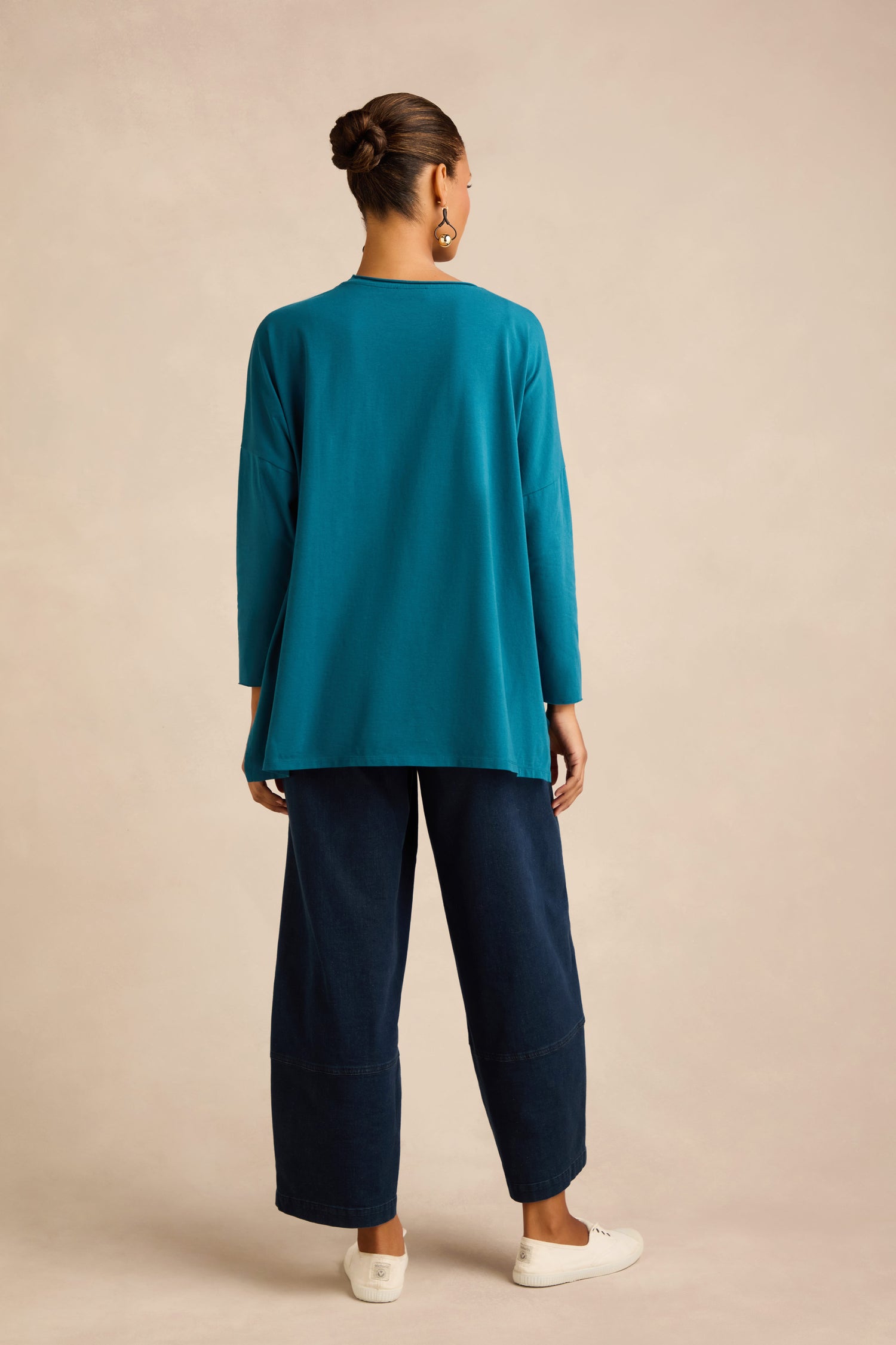 A woman stands facing away against a beige background, wearing the Cotton Jersey Relaxed Top in teal with a boxy silhouette, wide-leg navy pants, white slip-on shoes, and large earrings.