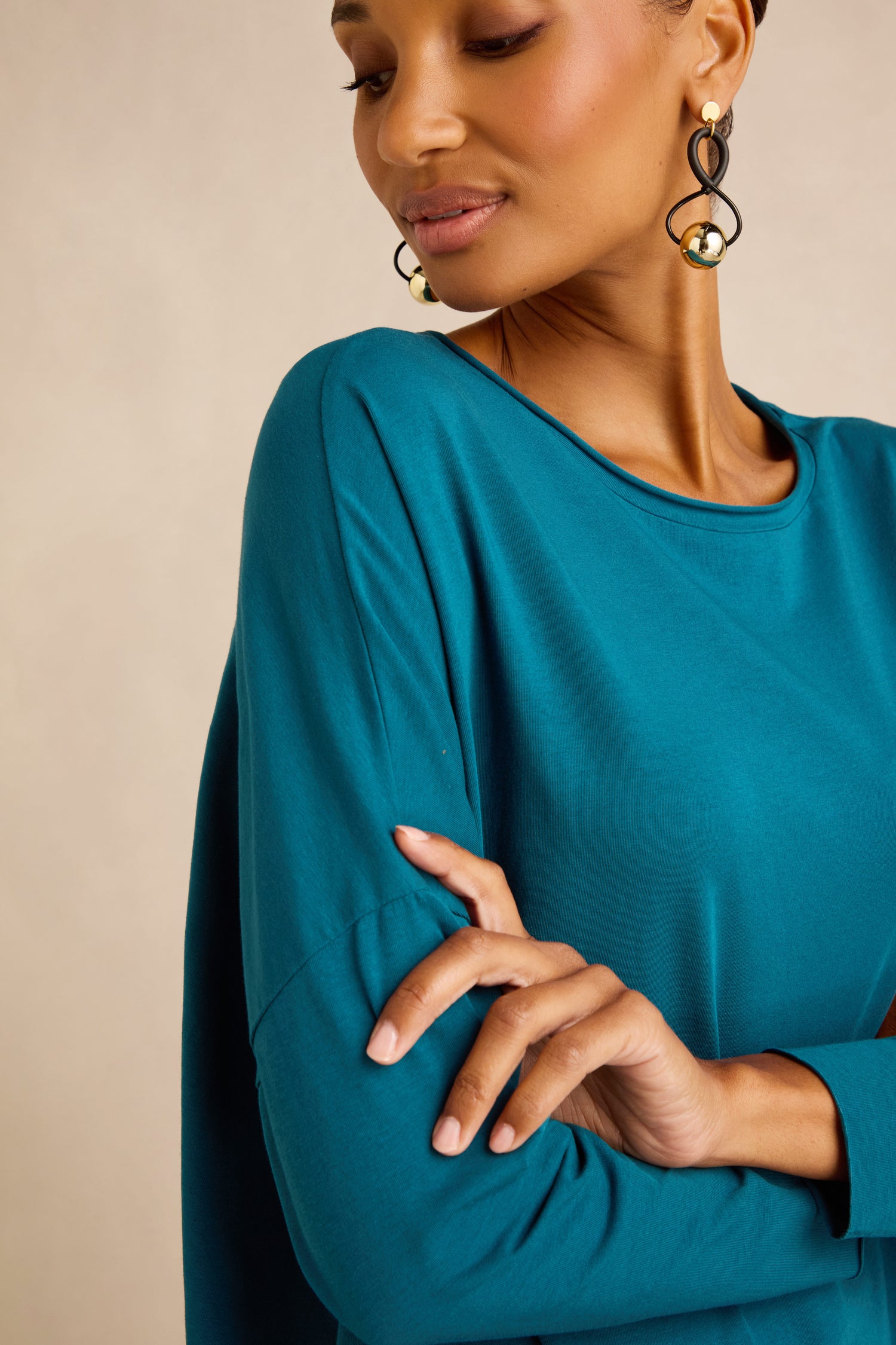 A woman models the Cotton Jersey Relaxed Top—a teal, long-sleeve style with a boxy silhouette in soft cotton jersey—accessorized with statement earrings as she looks down, one arm crossed and her hand resting on her upper arm.