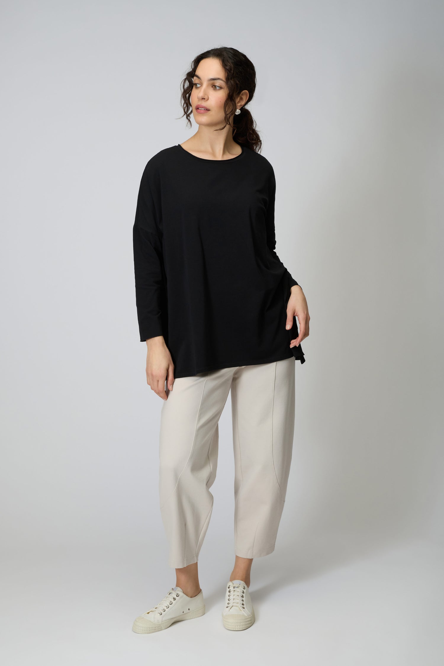 A person stands against a plain background wearing the Cotton Jersey Relaxed Top in black, paired with light cropped pants and white sneakers—a true wardrobe essential.