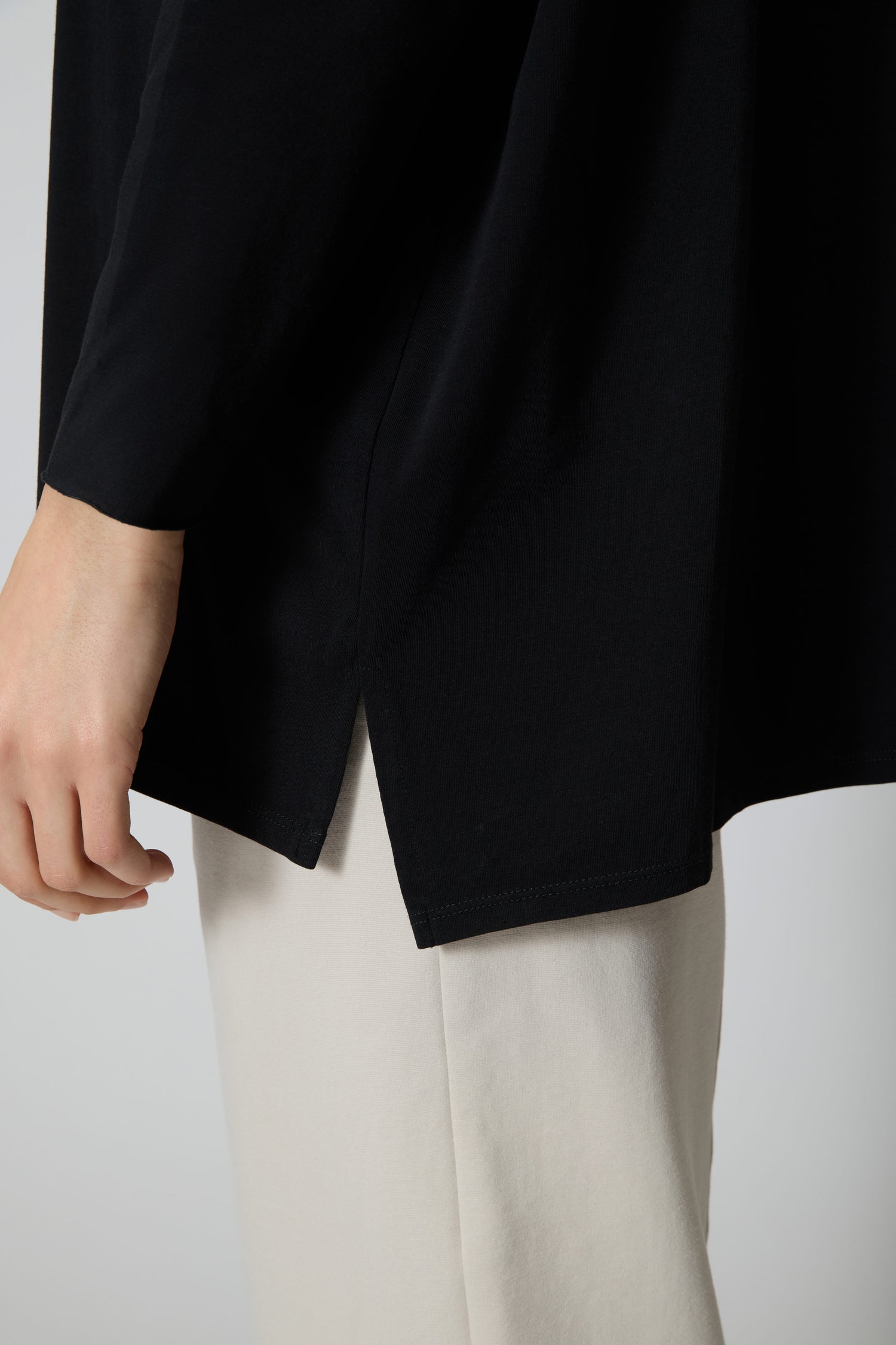A person wearing the Cotton Jersey Relaxed Top, a relaxed fit black long-sleeve with side slit, paired with light-colored pants, shown from the side at waist level.
