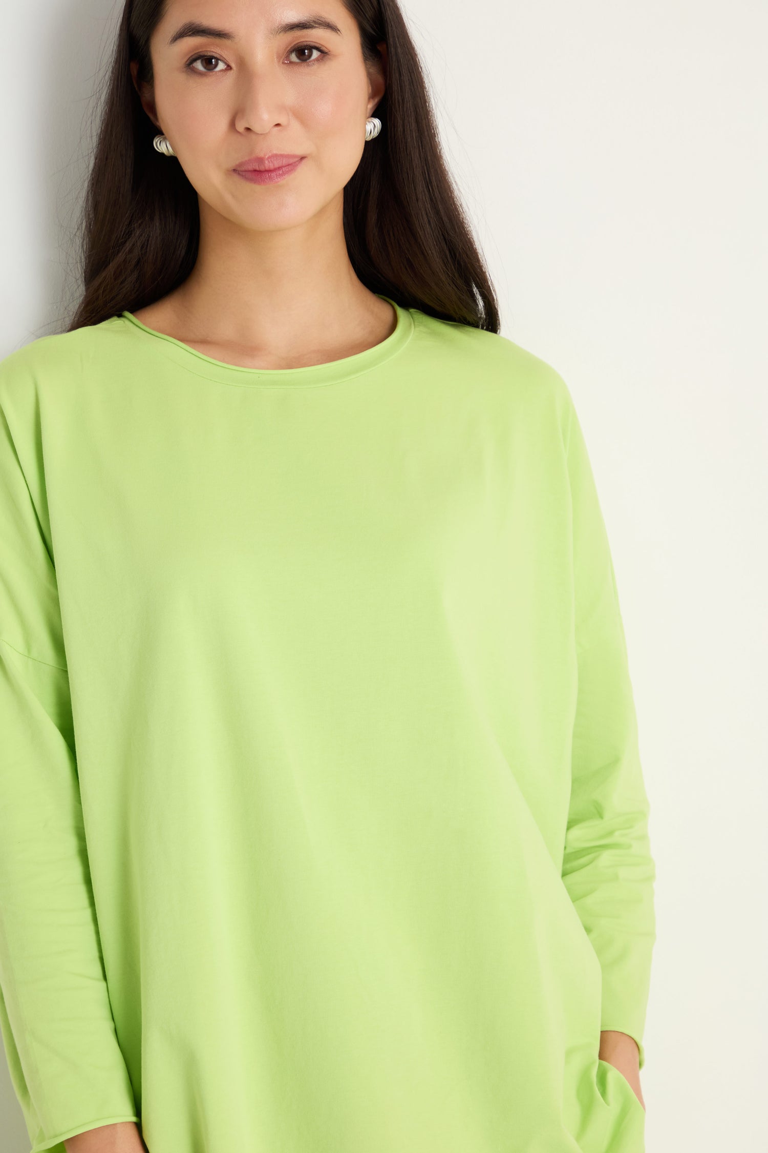 A woman with long dark hair wears the Cotton Jersey Relaxed Top in light green, featuring a relaxed silhouette. She stands against a white background and looks at the camera.