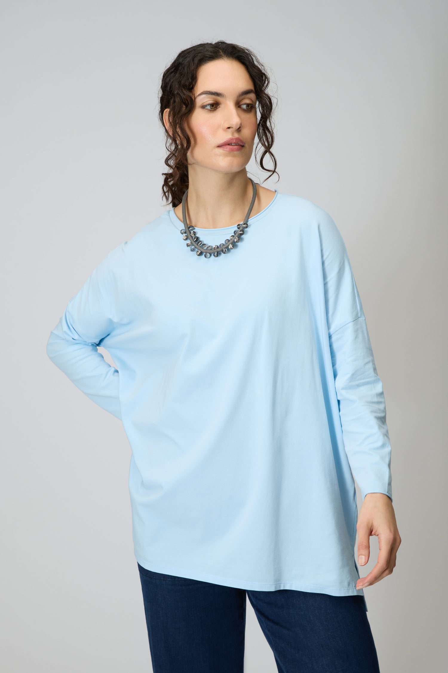 Woman wearing the Cotton Jersey Relaxed Top in light blue with dark blue jeans and a beaded necklace, standing against a plain, light background.