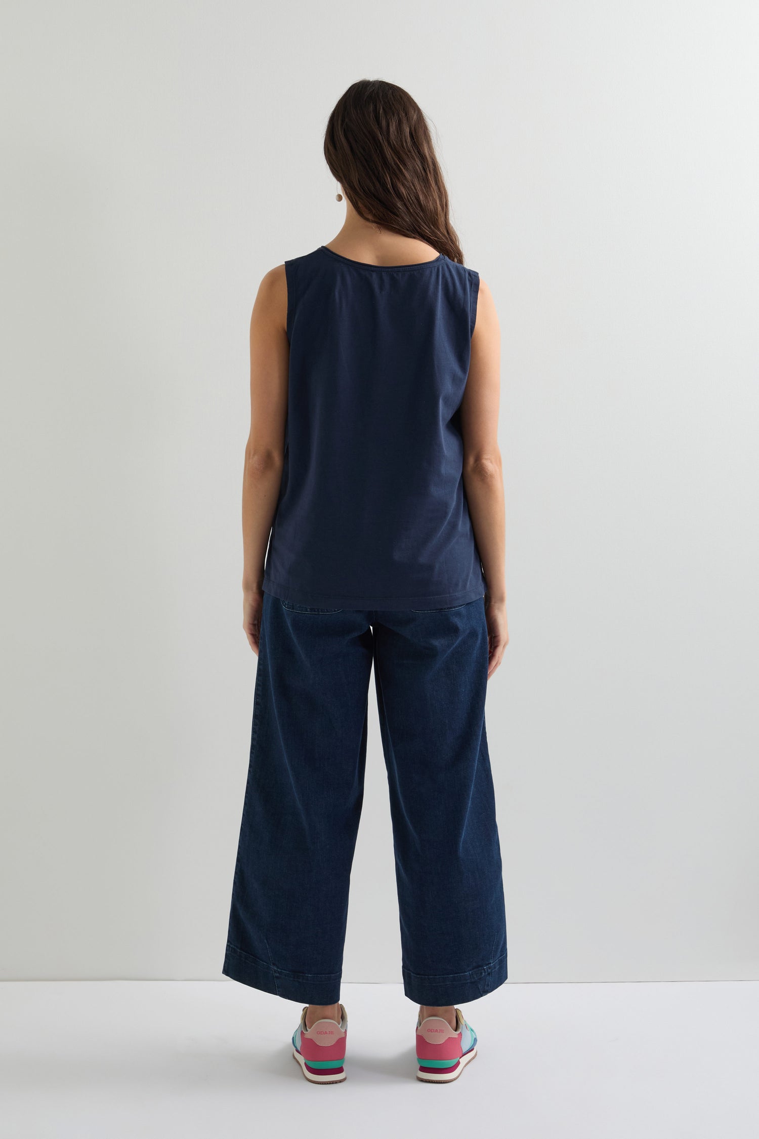 With their back to the camera, they showcase a relaxed fit in a navy Cotton Jersey Boxy Vest, paired with wide-leg jeans and colorful shoes, all against a plain white background.