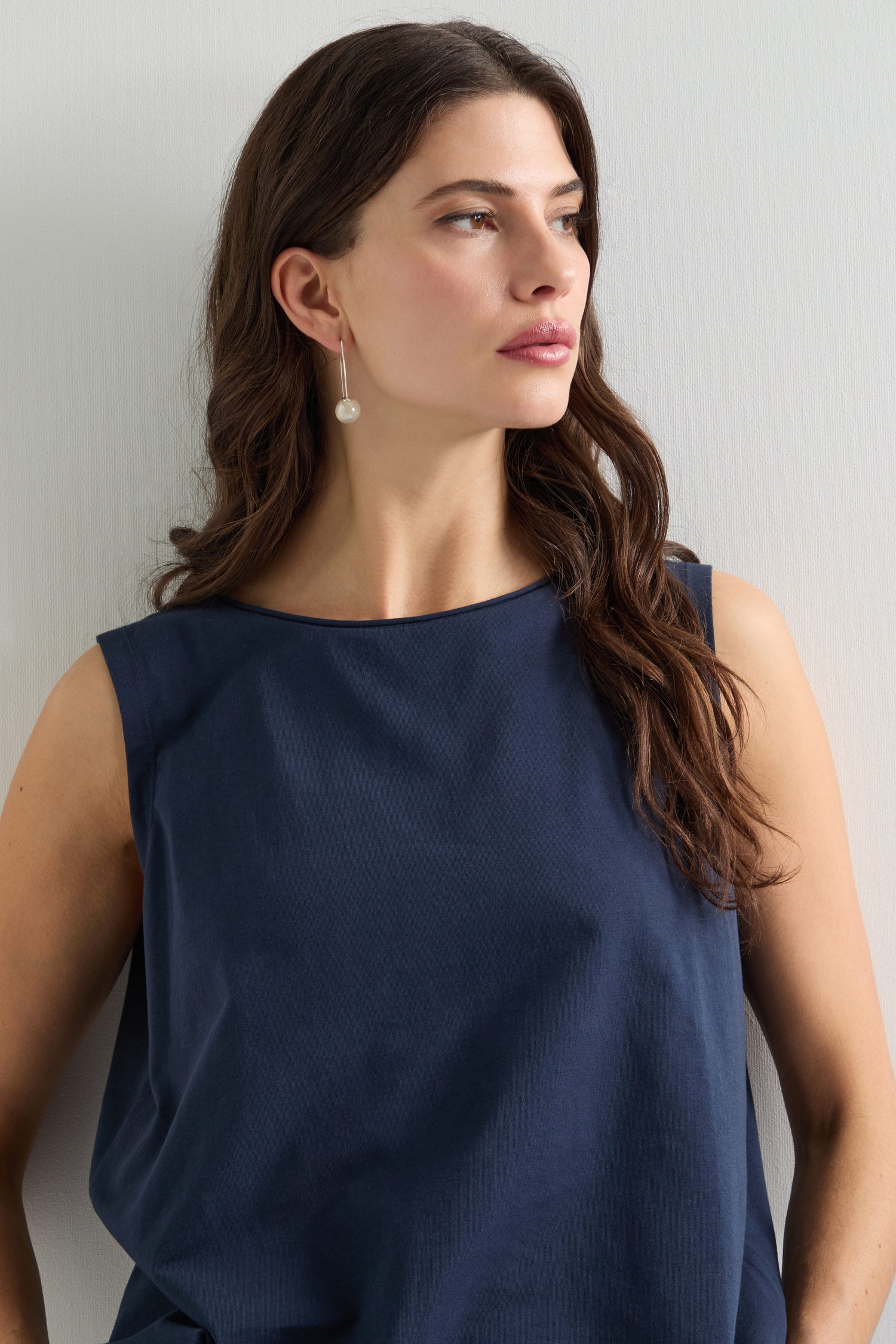 A person with long brown hair wears the Cotton Jersey Boxy Vest, made from breathable stretch jersey, and pearl drop earrings, gazing sideways against a plain background.
