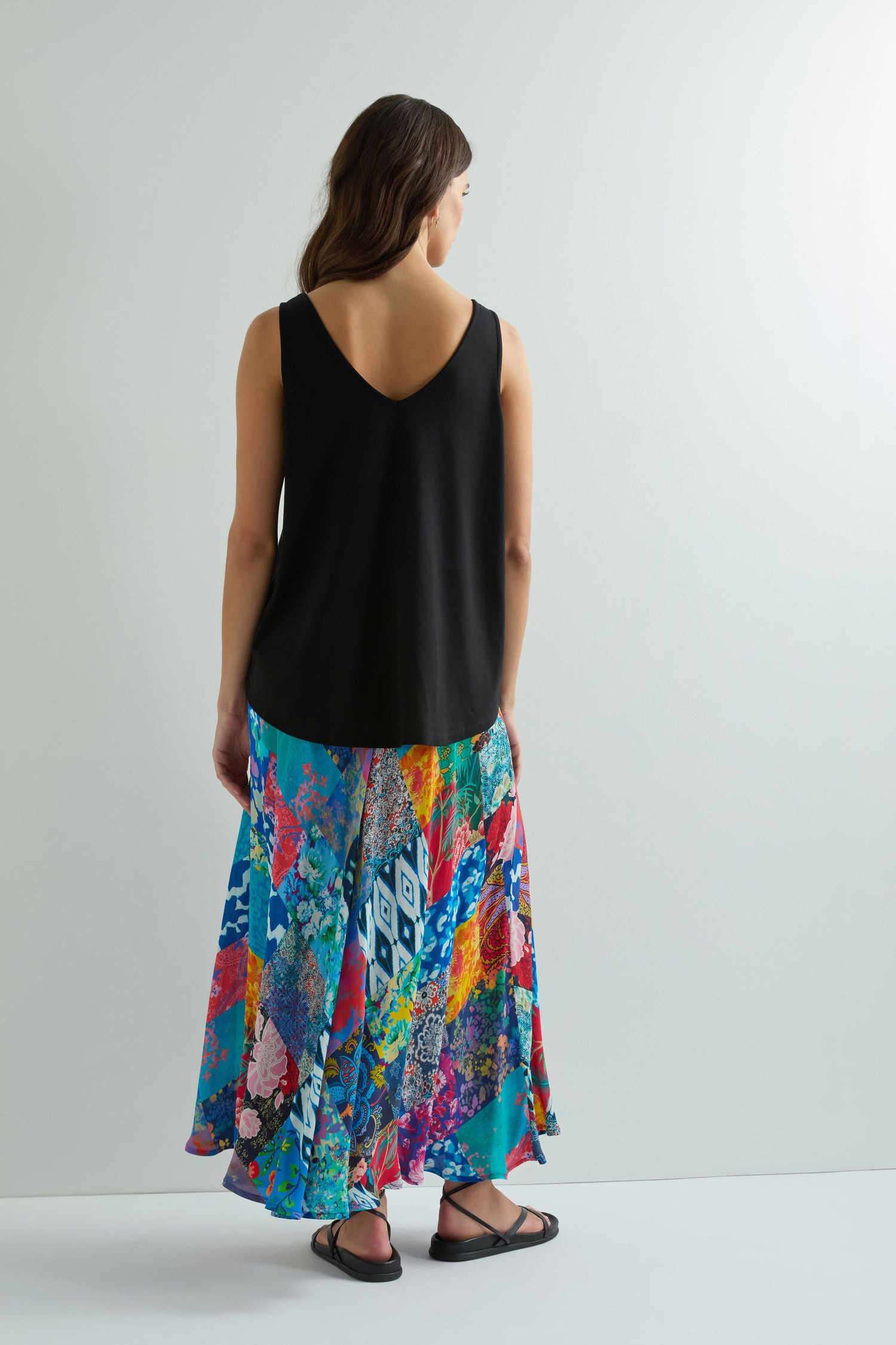 A woman faces away, wearing a Viscose Jersey V Neck Vest in black, a colorful floral and geometric print skirt, and black sandals against a plain white background.