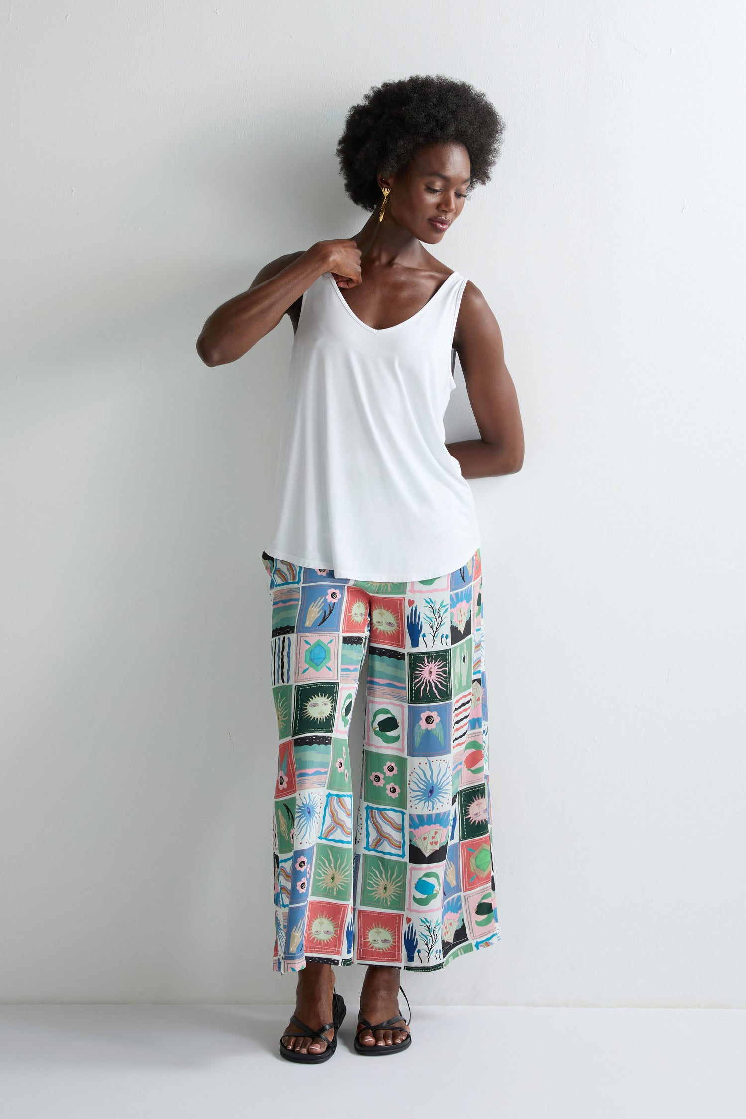 A woman stands against a white wall wearing the Viscose Jersey V Neck Vest, wide-leg patterned pants with colorful squares, and black sandals—a vibrant look for summer.
