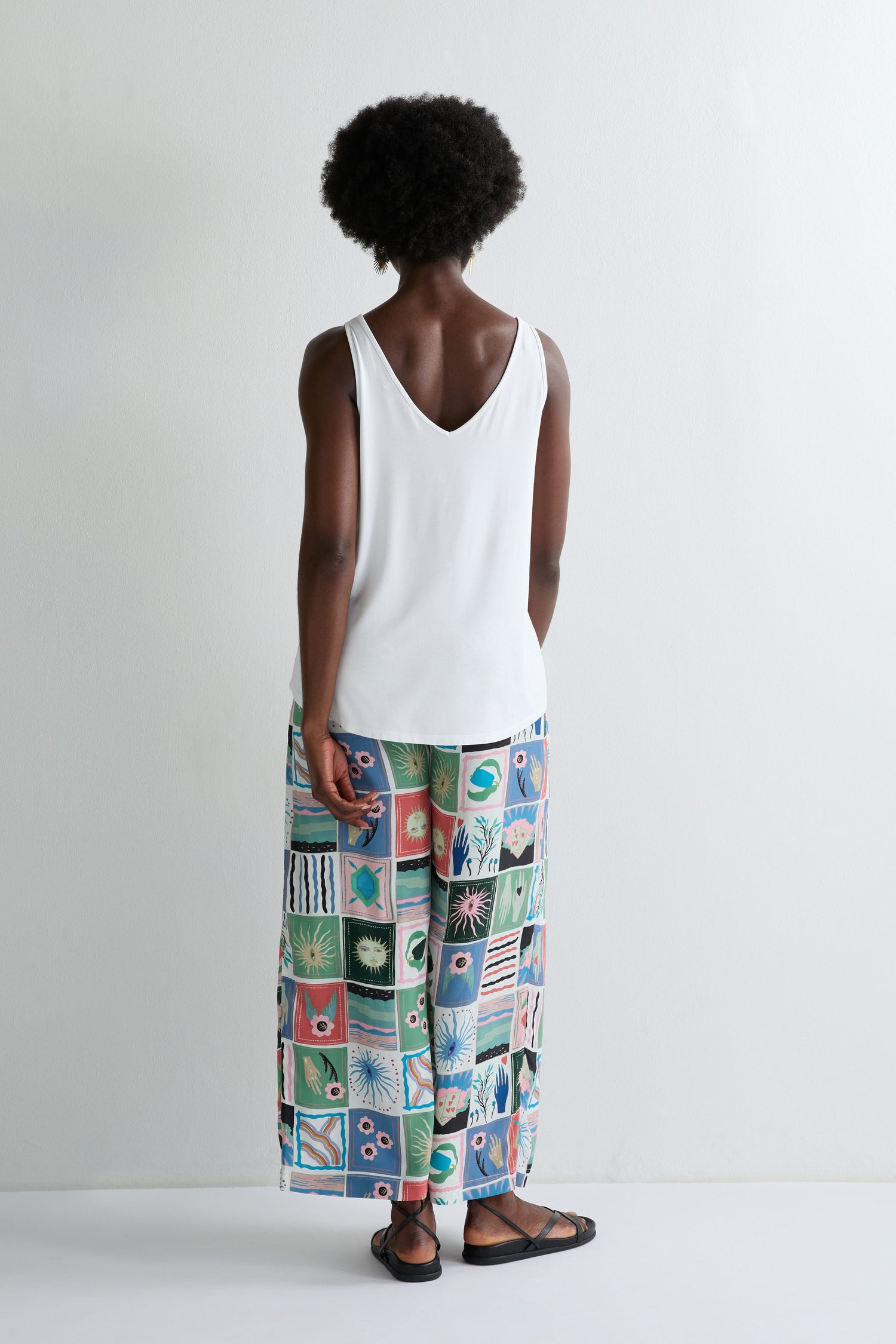A person stands facing away in a white Viscose Jersey V Neck Vest and colorful patterned wide-leg pants with black sandals, set against a plain white background—essential summer wardrobe pieces.