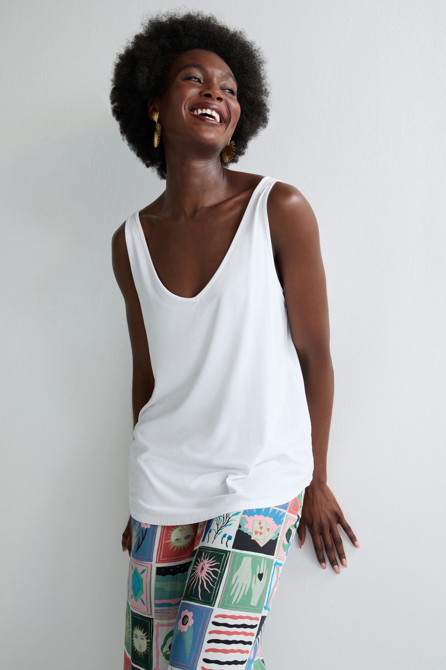 A woman in a white Viscose Jersey V Neck Vest and patterned pants stands against a white wall, smiling and looking to the side—making the Viscose Jersey V Neck Vest a must-have for any summer wardrobe.