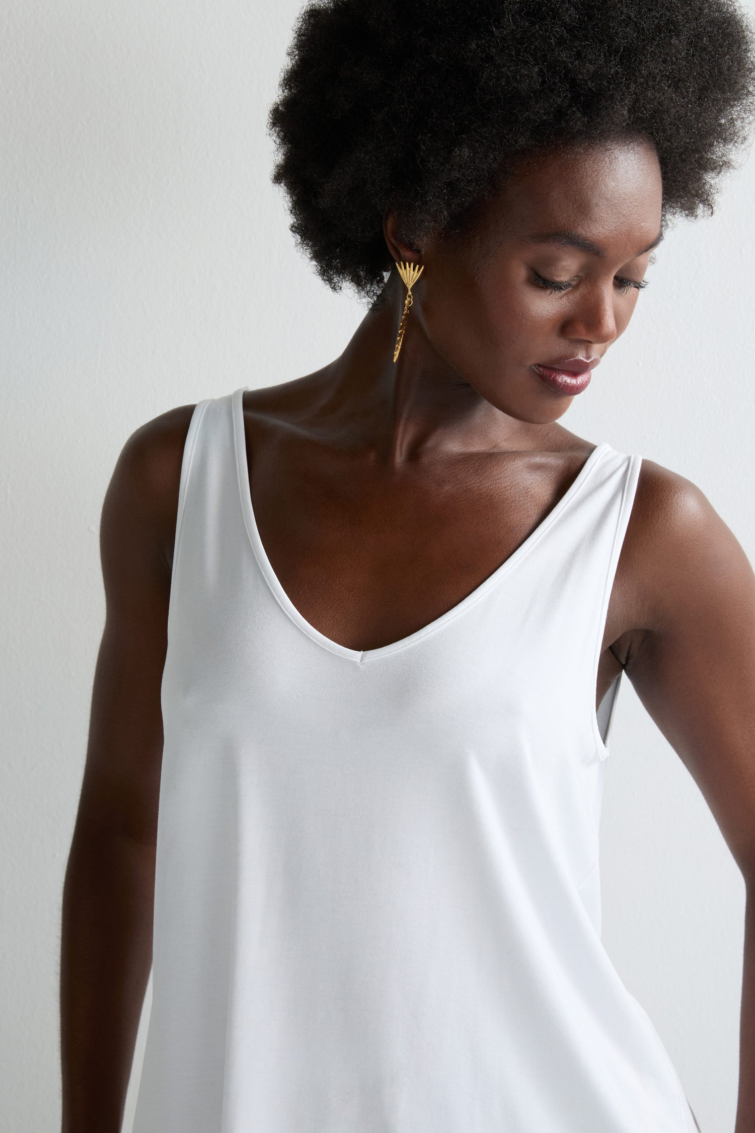 A woman with an afro hairstyle wears the Viscose Jersey V Neck Vest in white and gold feather-shaped earrings, looking downwards against a plain light background.