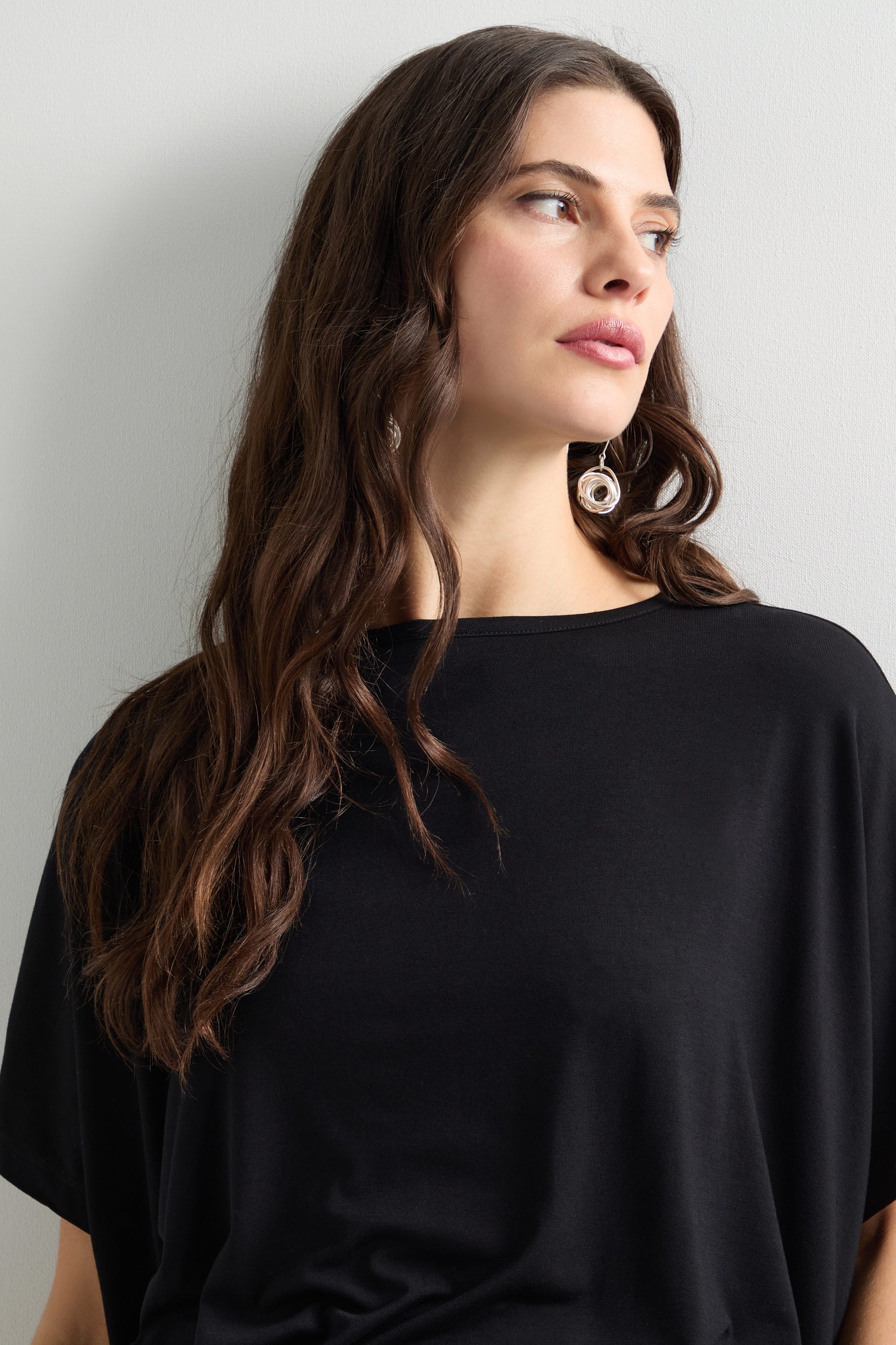 A woman with long brown hair dons a Viscose Jersey Square Top and large round earrings, gazing to the side against a plain backdrop, capturing the essence of everyday dressing.