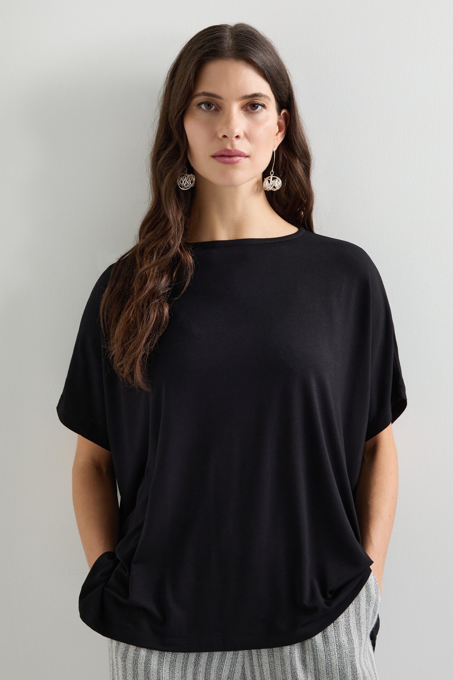 A person with long hair wearing a black Viscose Jersey Square Top and striped pants stands against a plain wall, hands in pockets, accentuated by silver earrings—ideal for an everyday look.