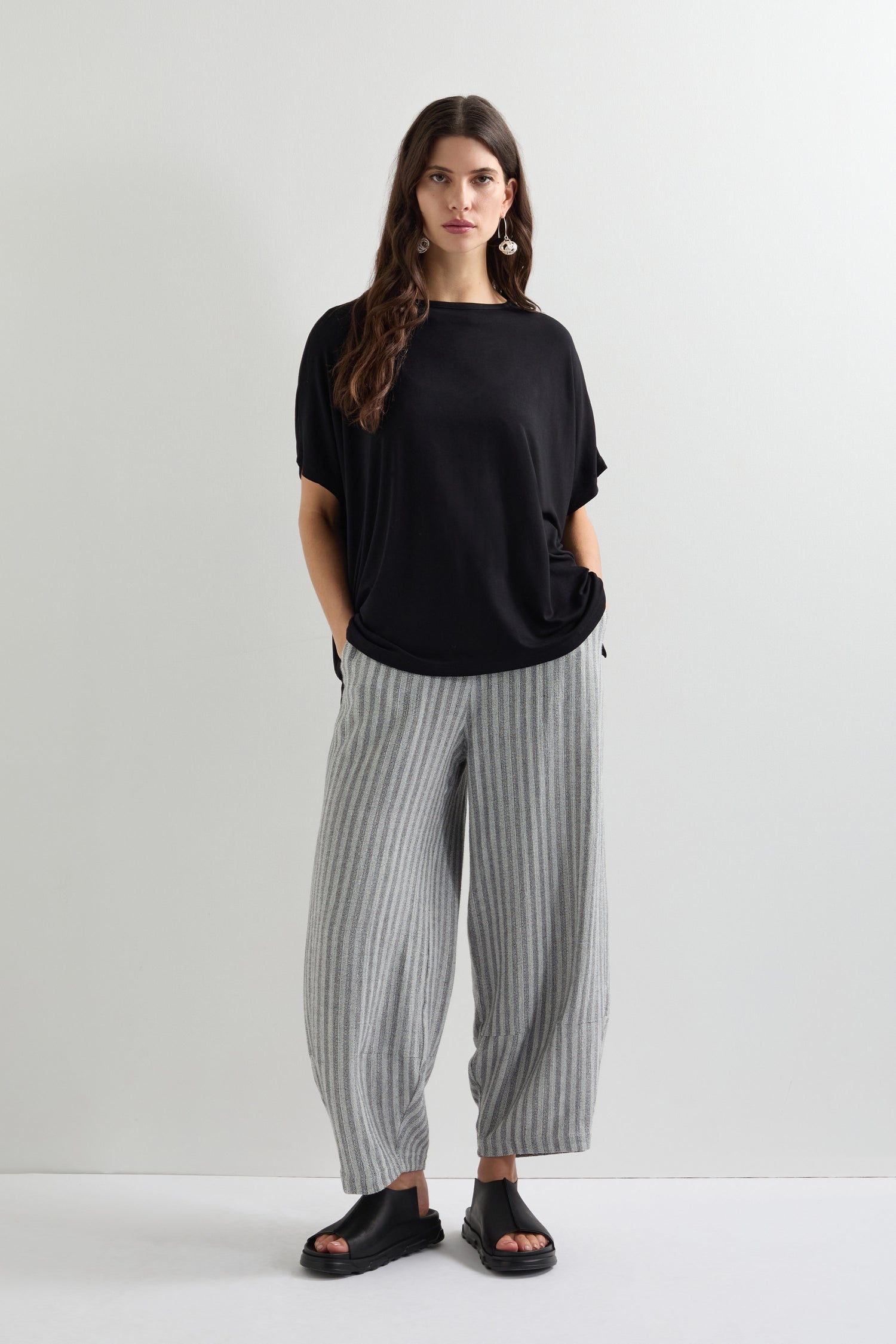 A person stands against a plain backdrop, hands in pockets, wearing a Viscose Jersey Square Top in black viscose jersey, complemented by gray striped pants and black sandals.