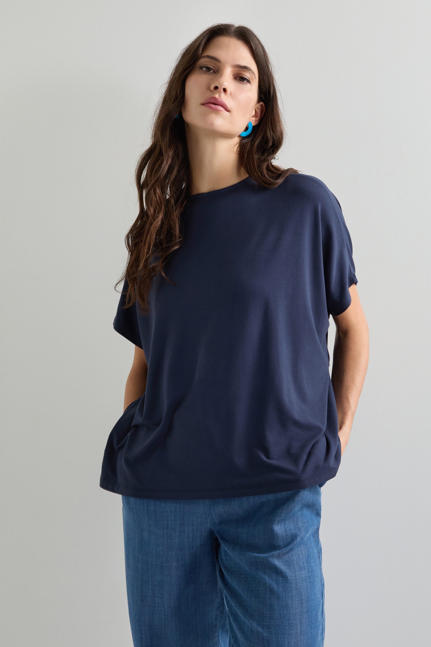 A person models a chic navy Viscose Jersey Square Top with blue pants, hands tucked in pockets against a plain backdrop—an ideal look for everyday dressing.
