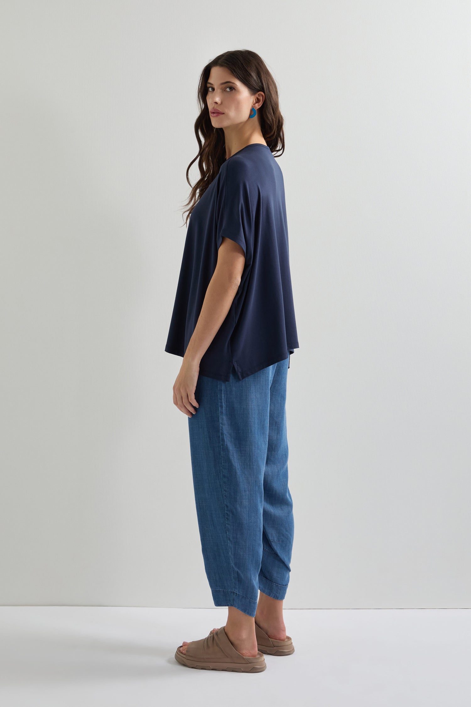A long-haired woman stands sideways wearing a loose navy Viscose Jersey Square Top, light blue pants, and brown sandals, embodying effortless everyday style against a white backdrop.