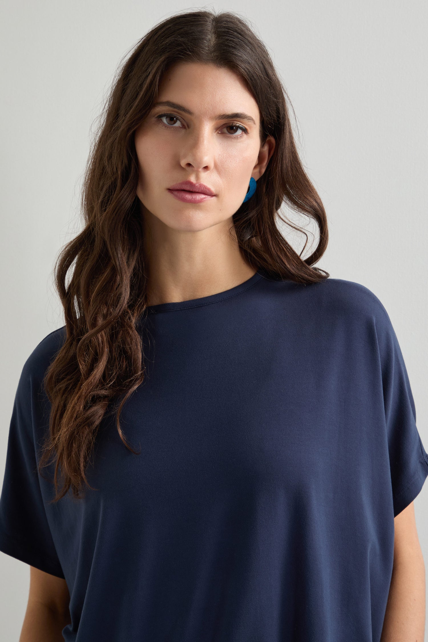 A long-haired individual with dark hair is against a plain white backdrop, styled in a Viscose Jersey Square Top in navy blue and blue earrings, embodying the essence of everyday fashion.