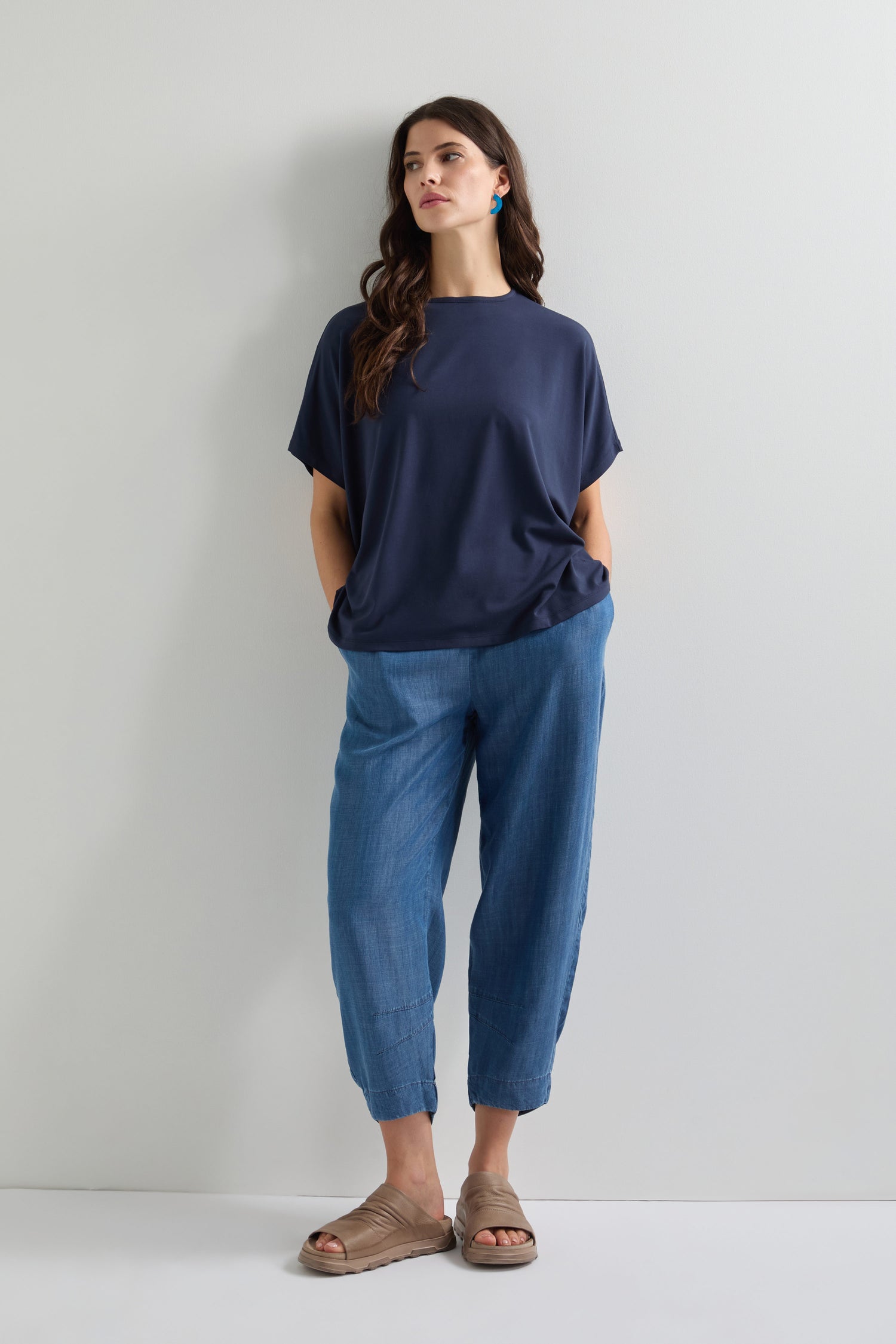 Wearing a Viscose Jersey Square Top, light blue pants, and beige sandals, someone stands casually against a light gray wall. Hands in pockets and glancing aside, their ensemble is the epitome of comfort and effortless style.