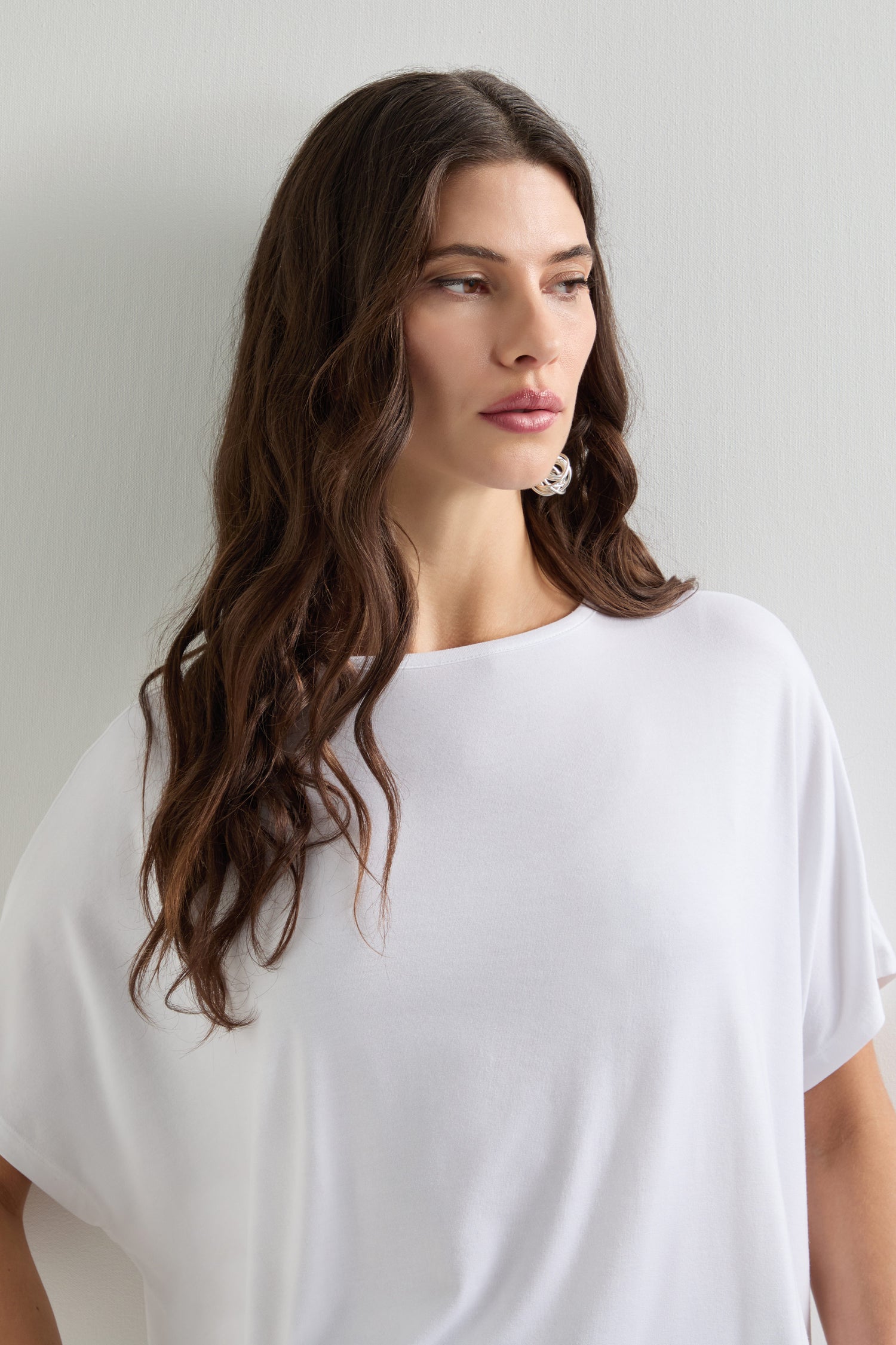 A woman with long brown hair stands against a neutral backdrop, wearing the Viscose Jersey Square Top, embodying effortless everyday style as she gazes to the side.
