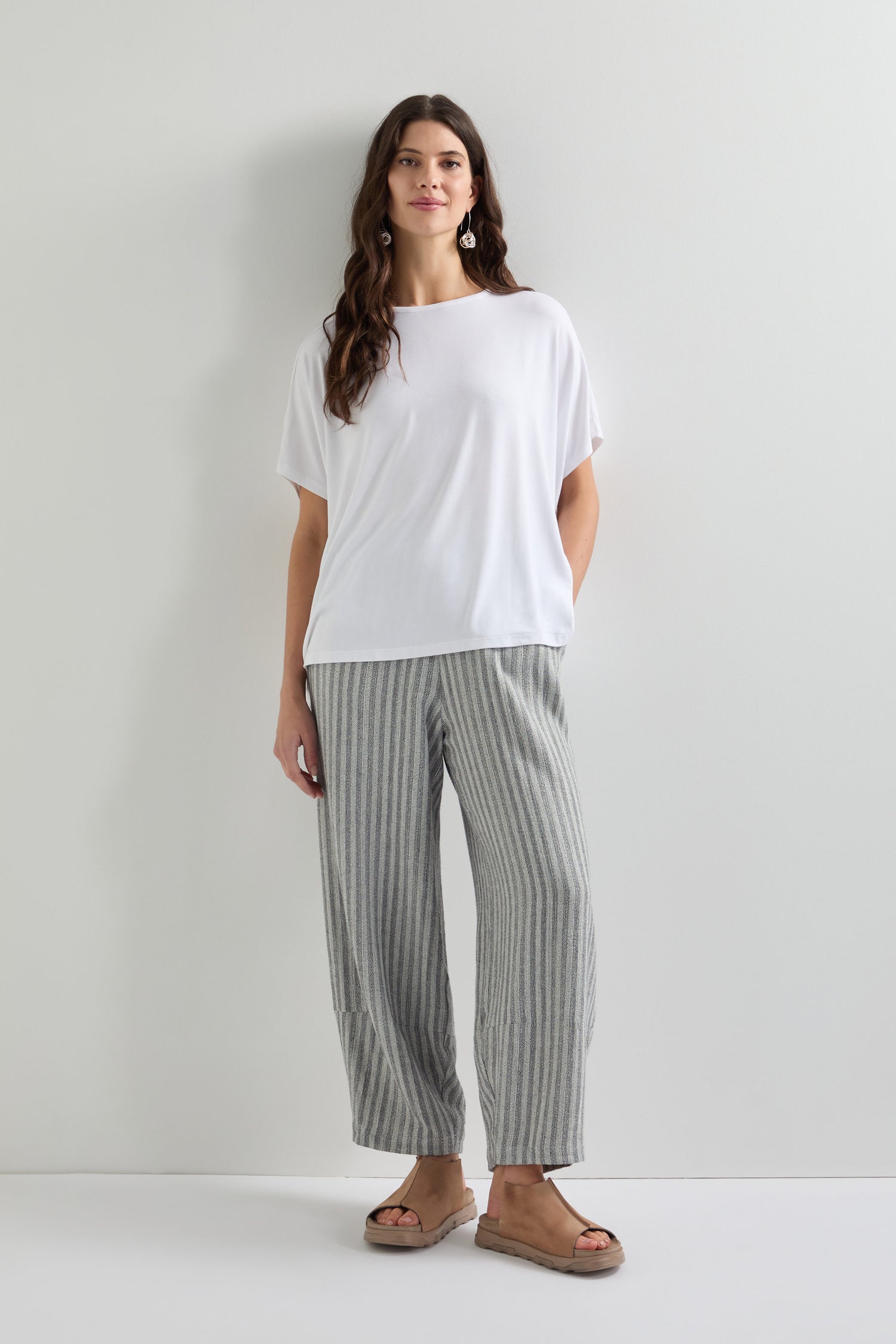 A person stands against a white wall, embracing everyday style in a Viscose Jersey Square Top, paired with gray striped pants and sandals.