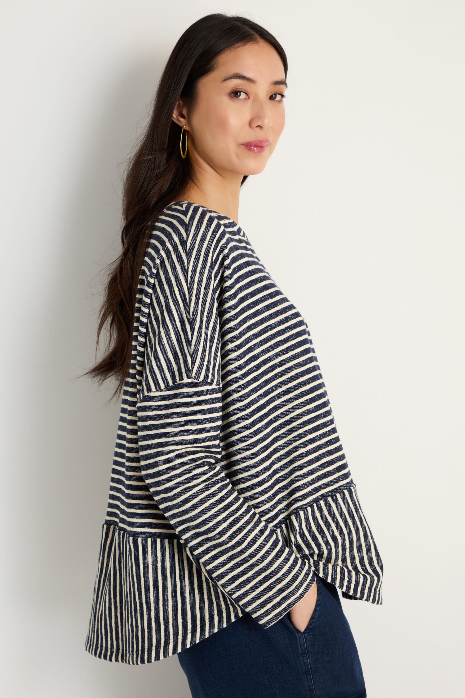 A woman stands sideways against a white wall, wearing the Textured Stripe Jersey Boxy Top in navy and white with dark jeans and her hands in her pockets.