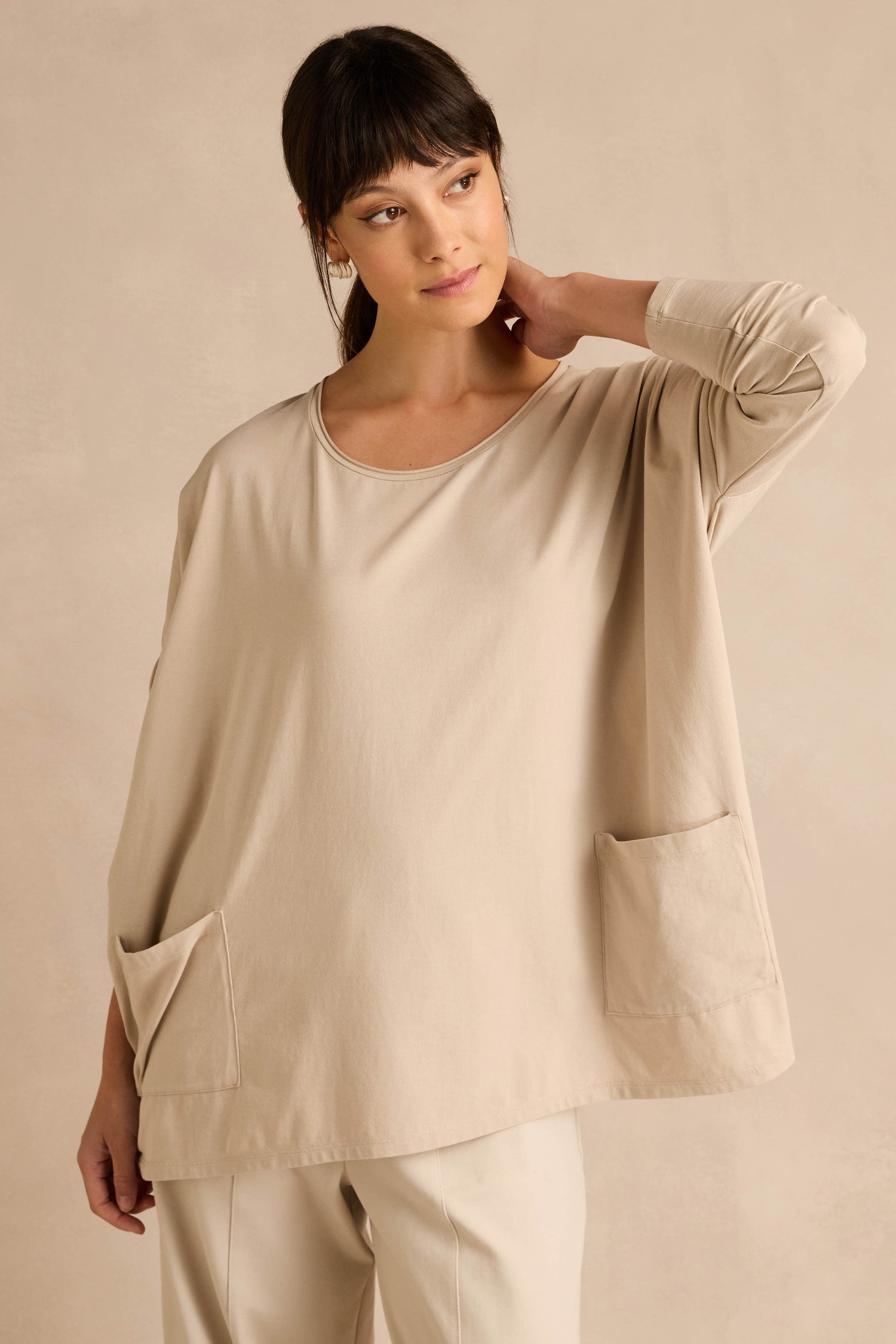 A woman wears the Ecru Cotton Jersey Pocket Top—an oversized, relaxed shirt in beige with two front pockets—paired with matching beige pants, standing against a beige wall with her hand resting on her neck.