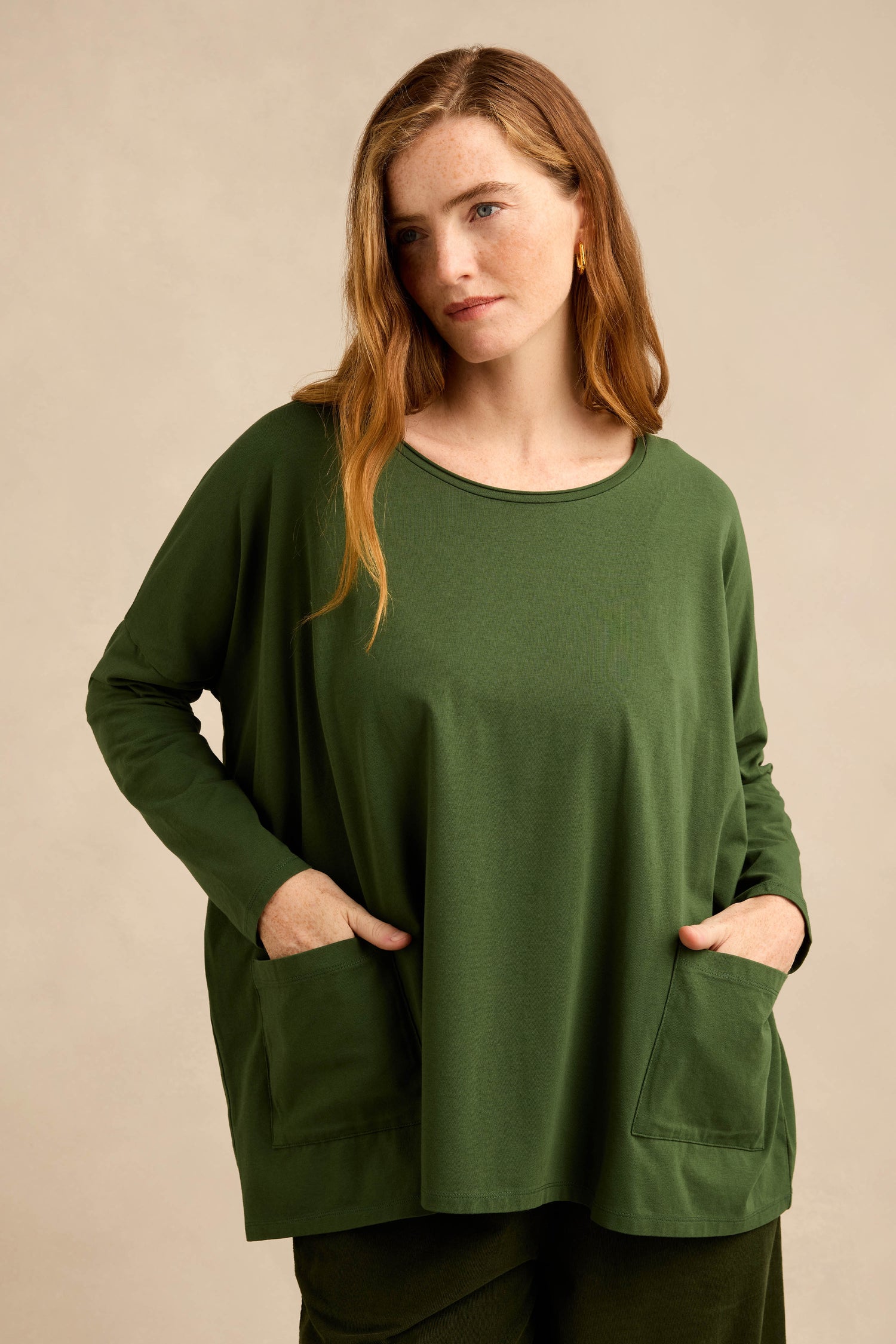 A woman stands against a plain beige background with her hands in the pockets of the Forest Cotton Jersey Pocket Top—a loose-fitting, boxy green top featuring front pockets.