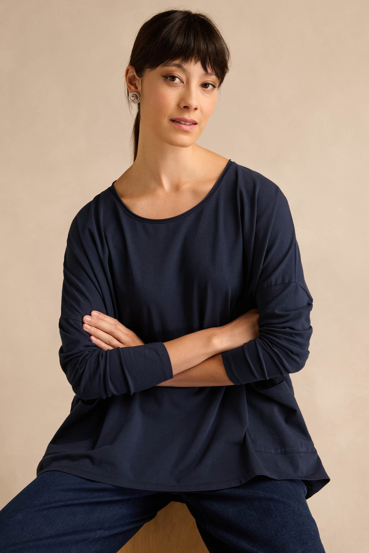 A woman with straight dark hair, wearing the Navy Cotton Jersey Pocket Top and jeans, sits with her arms crossed against a plain beige background.
