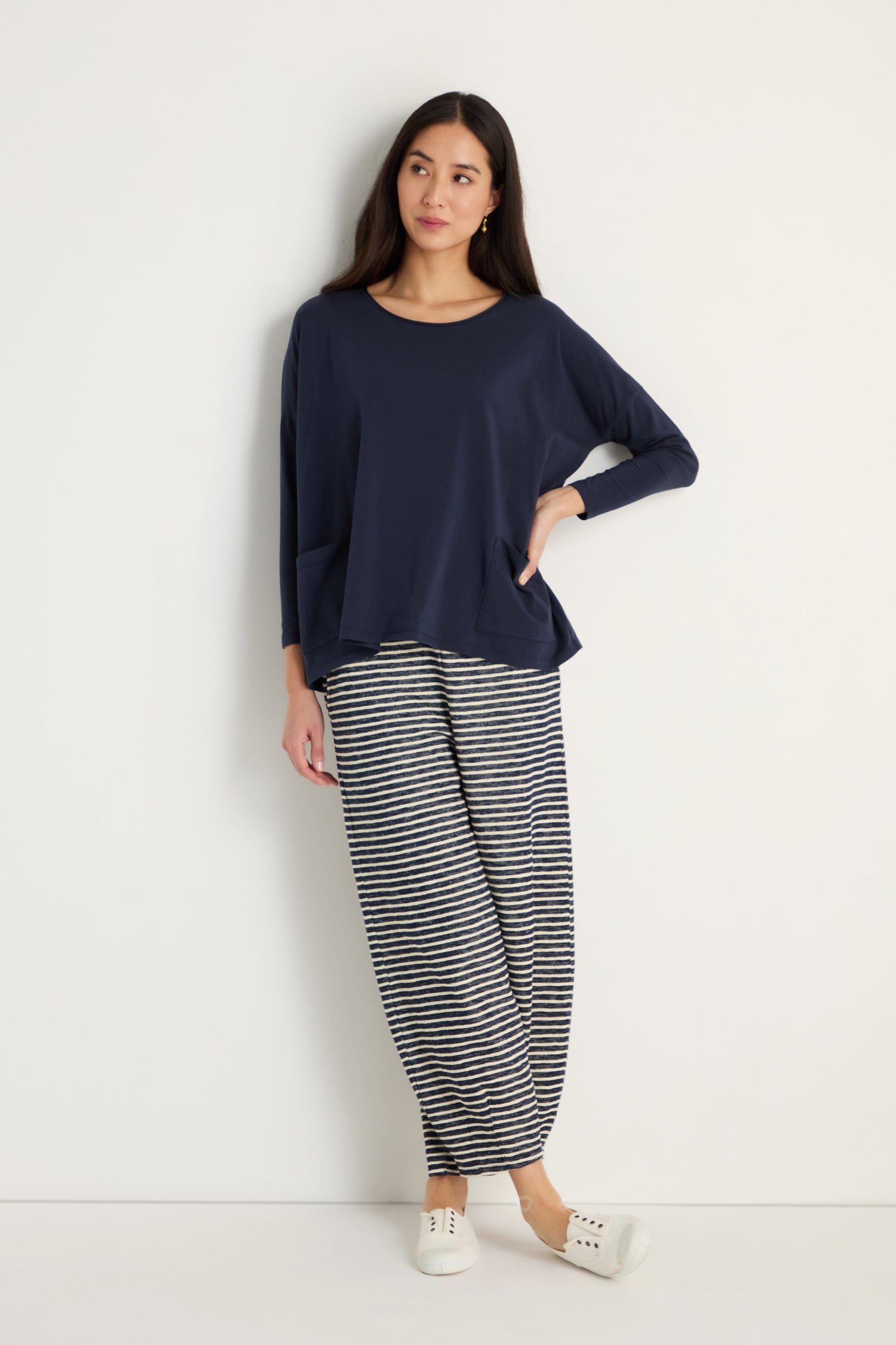 A woman stands against a plain white wall, wearing the Cotton Jersey Boxy Pocket Top in navy, styled with striped wide-leg pants and white slip-on shoes.