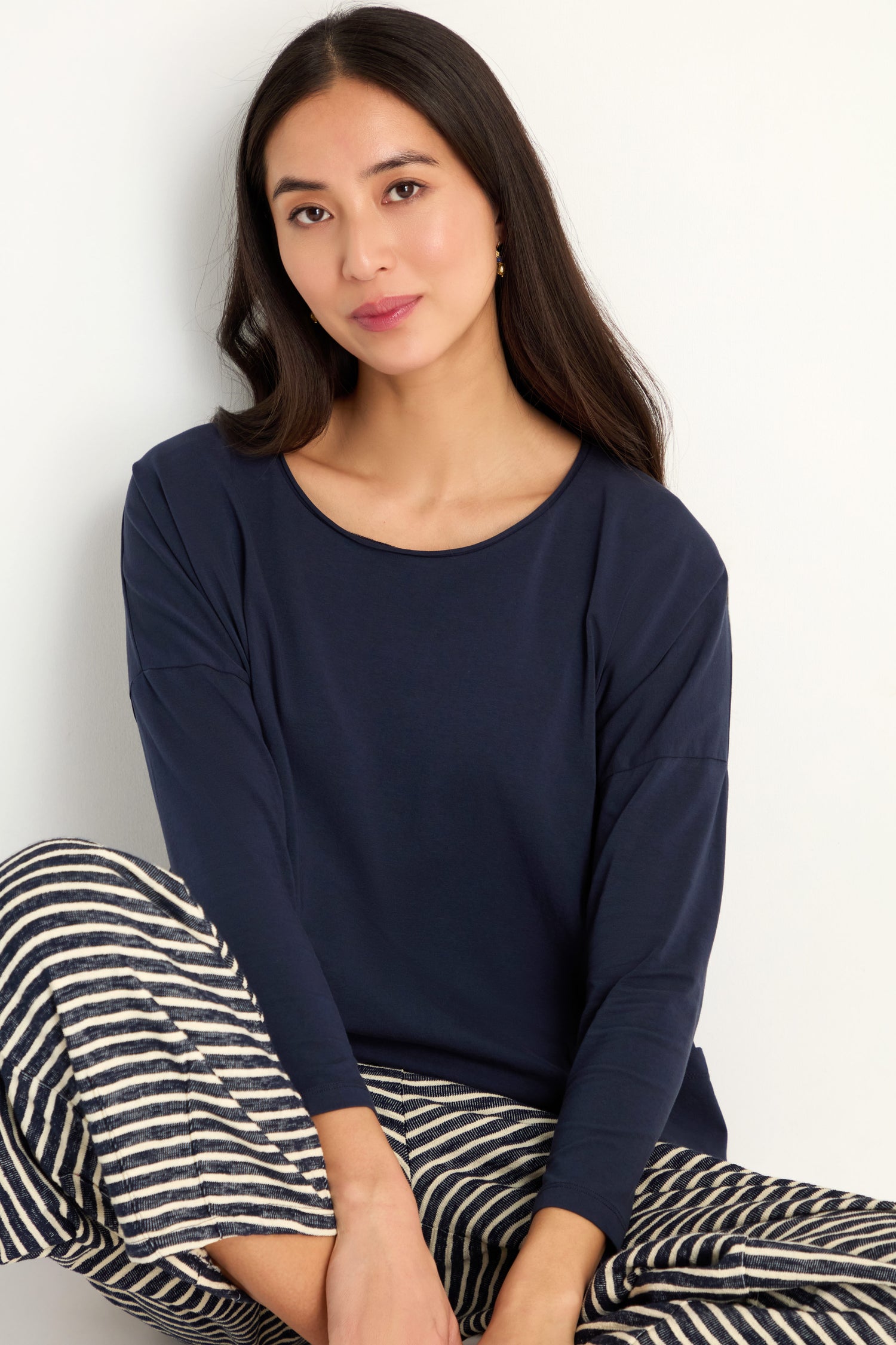 Woman with long dark hair wearing an oversized fit navy Cotton Jersey Boxy Pocket Top and black-and-white striped pants, sitting against a white background.