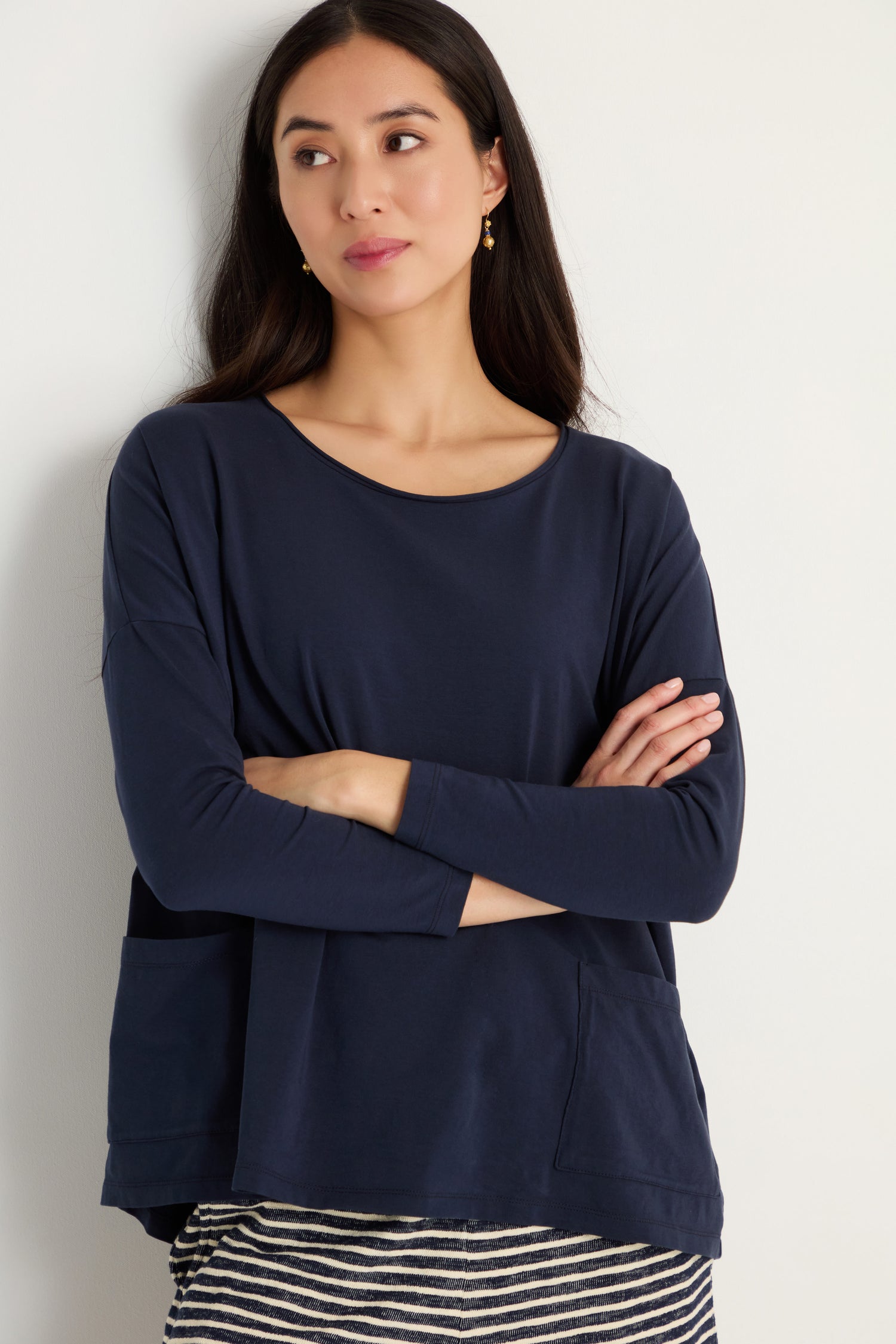 A woman with long dark hair stands against a light wall, arms crossed, wearing the Cotton Jersey Boxy Pocket Top in navy blue with an oversized fit and a striped skirt.