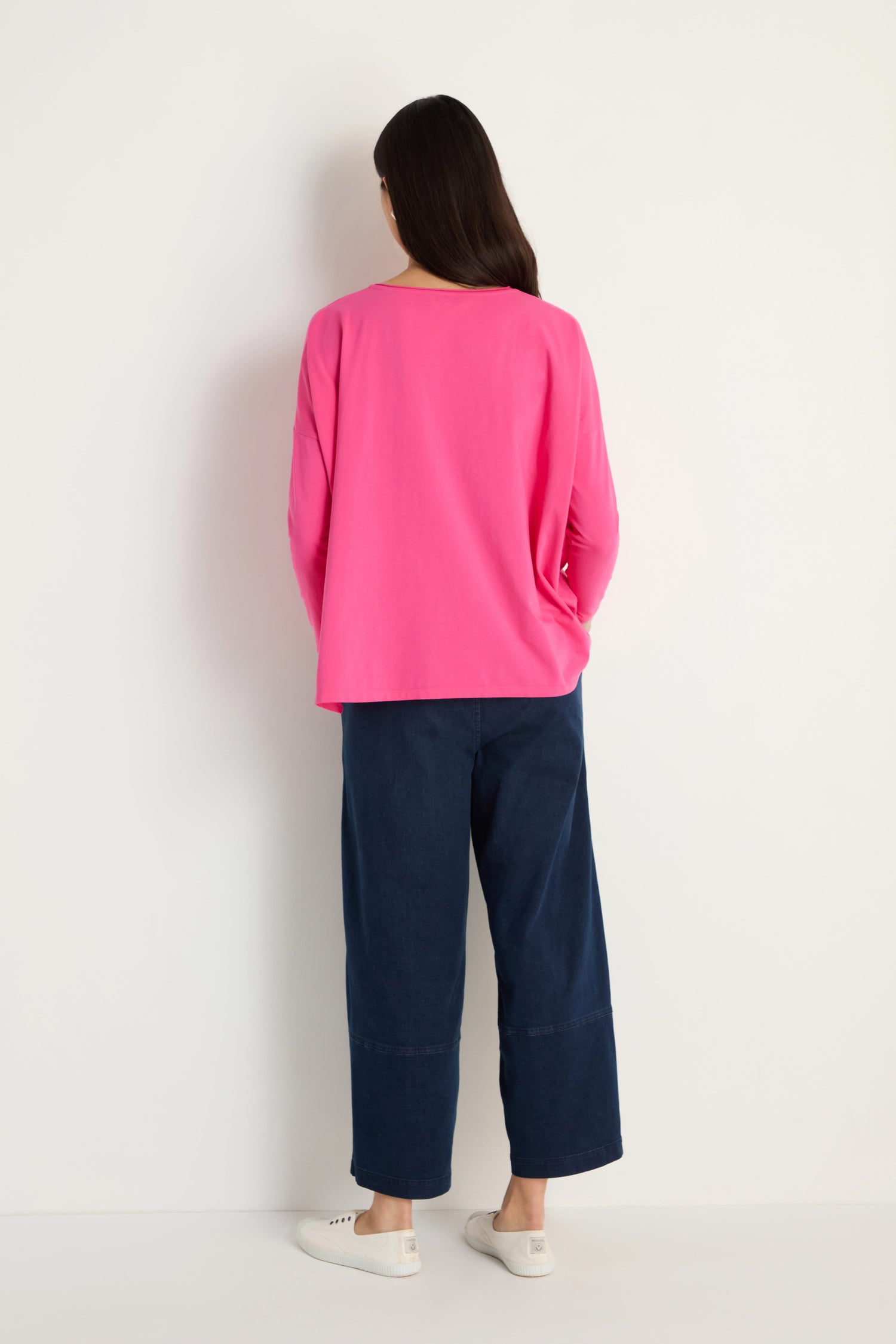 A person stands facing a white wall, wearing a bright pink Cotton Jersey Boxy Pocket Top, wide-leg dark blue jeans, and white sneakers.