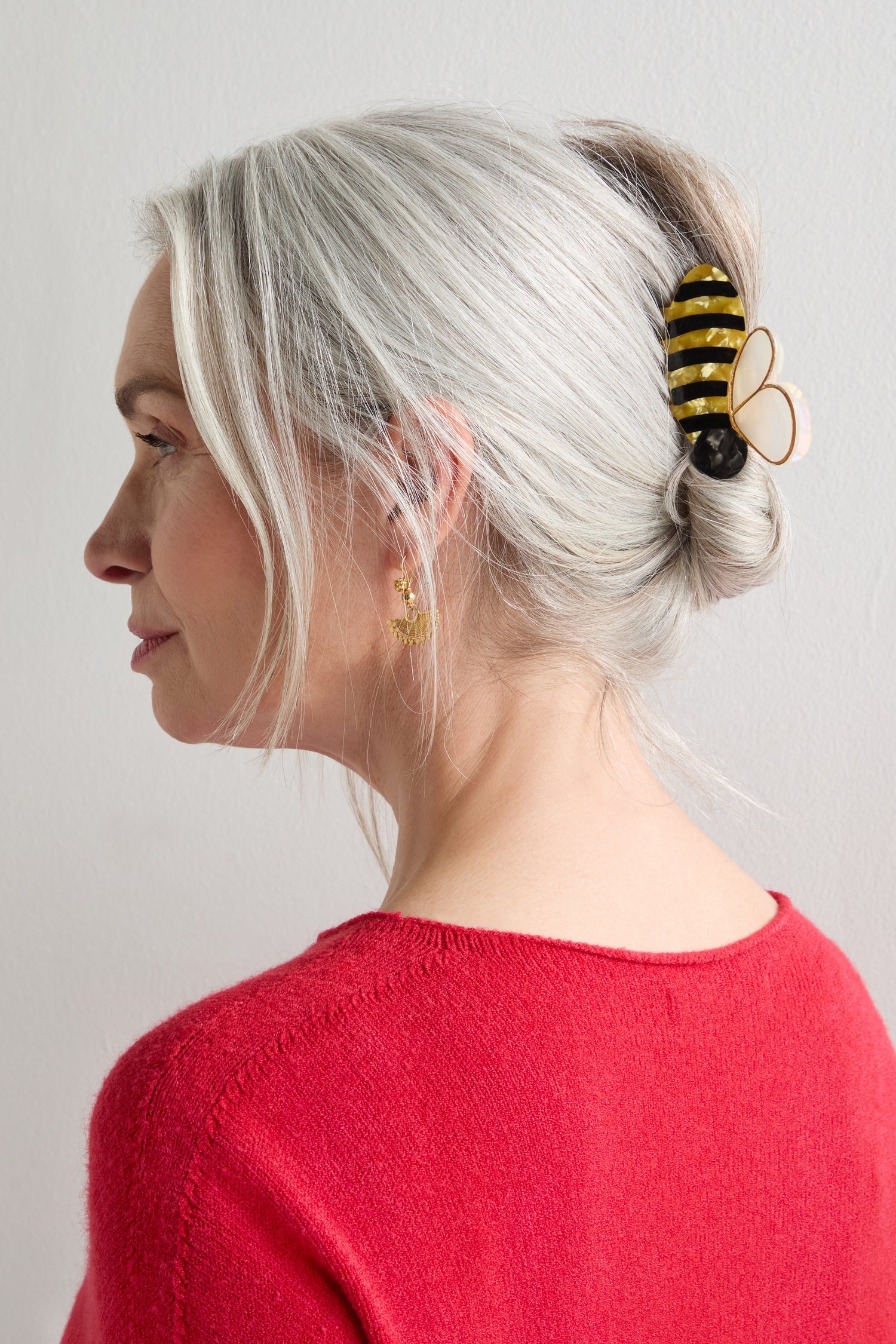 A woman with gray hair in a twist, secured with a Hand-Painted Bee Hair Clip, wears a red sweater and gold earrings, facing left against a plain light background.