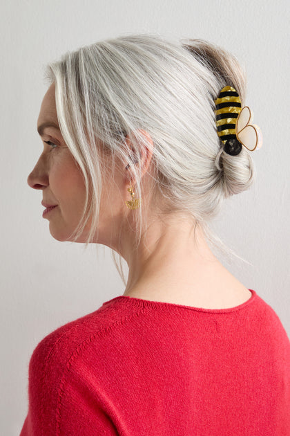 A woman with gray hair in a twist, secured with a Hand-Painted Bee Hair Clip, wears a red sweater and gold earrings, facing left against a plain light background.