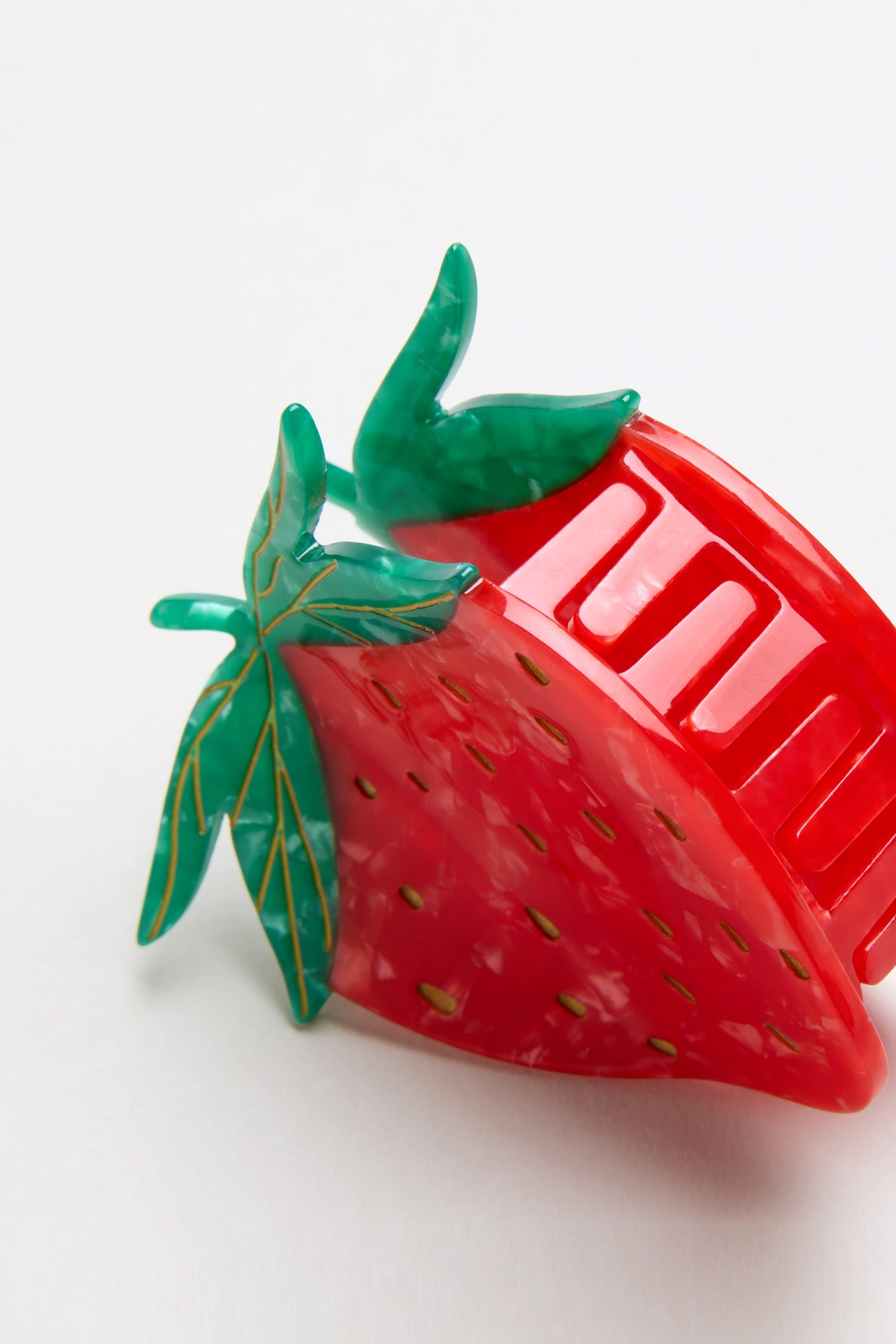 The Hand Painted Strawberry Hair Clip features eco-friendly materials, a red and green strawberry design with textured leaves and yellow seed details, displayed on a plain white background.