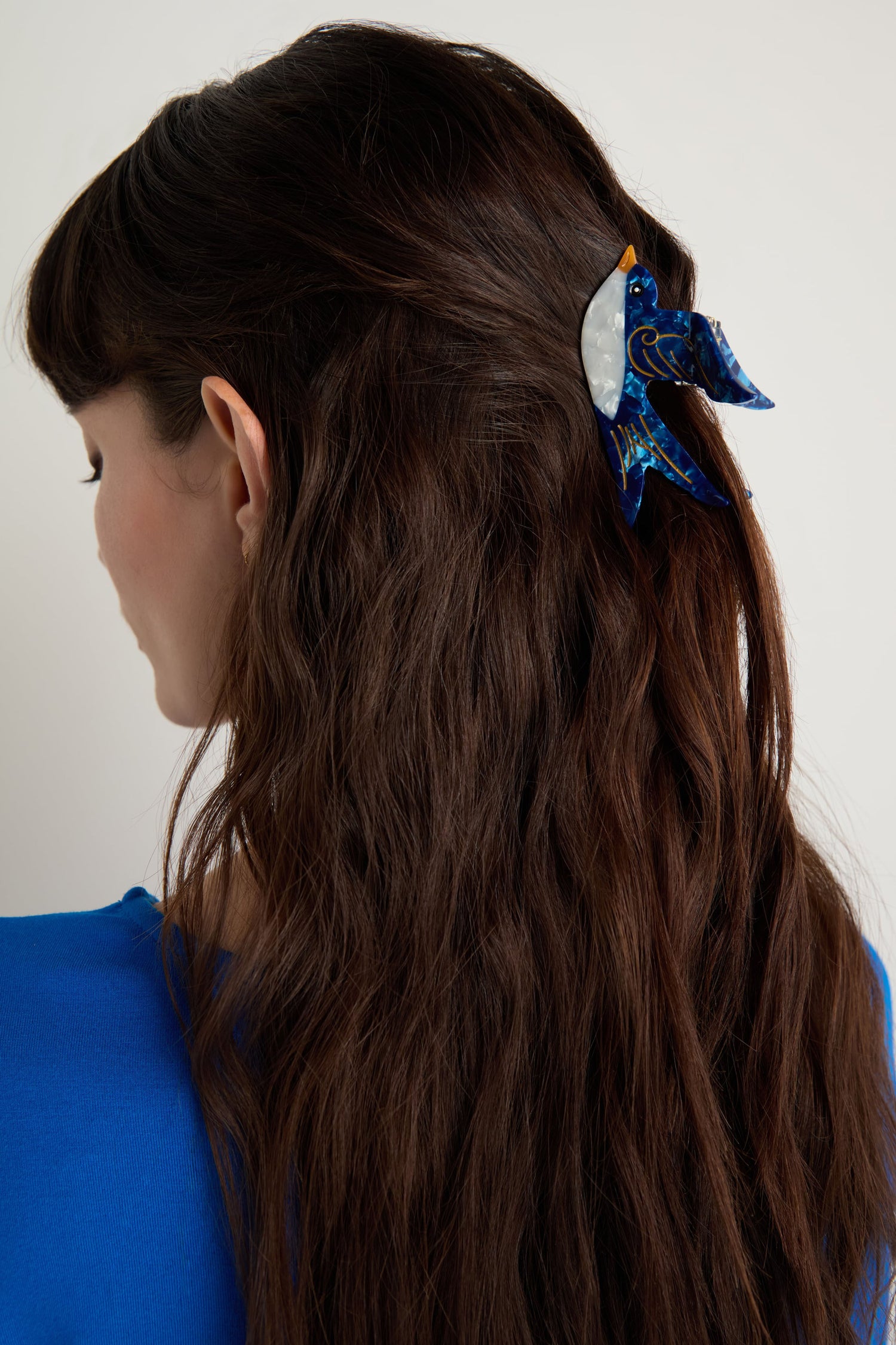 A woman with long, wavy brown hair is seen from behind, wearing a blue top and a hand-painted Swallow Hair Clip—an eco-friendly accessory featuring a decorative crescent moon detail.