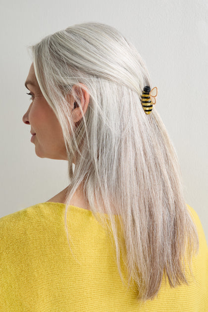 A woman with long gray hair, shown from the side against a light background, styles her hair with a Hand-Painted Mini Bee Hair Clip featuring opalescent wings. She is wearing a yellow top.