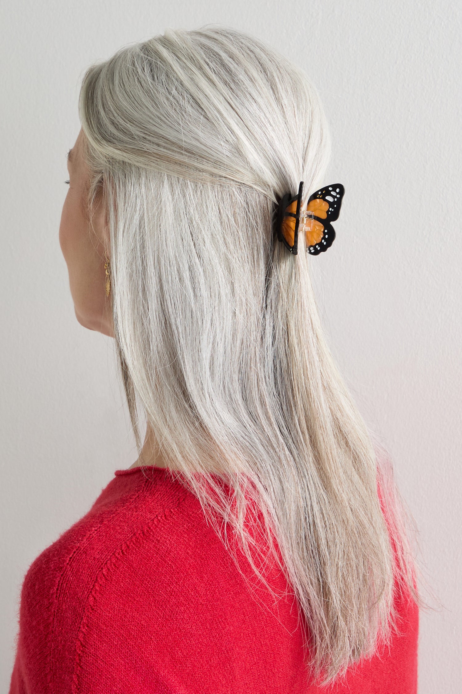 A person with long gray hair styled half up, secured with a Hand-Painted Mini Monarch Butterfly Hair Clip, wearing a red sweater and shown from the back.