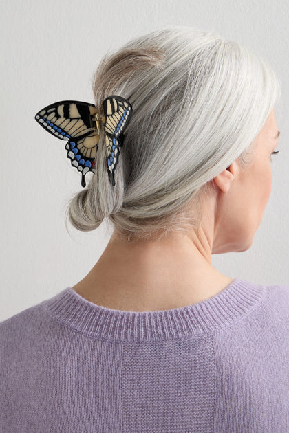 A person with gray hair in a low bun, secured by a Hand-Painted Swallowtail Hair Clip, gold-tone and butterfly-shaped, wears a lavender sweater and is shown from the back.