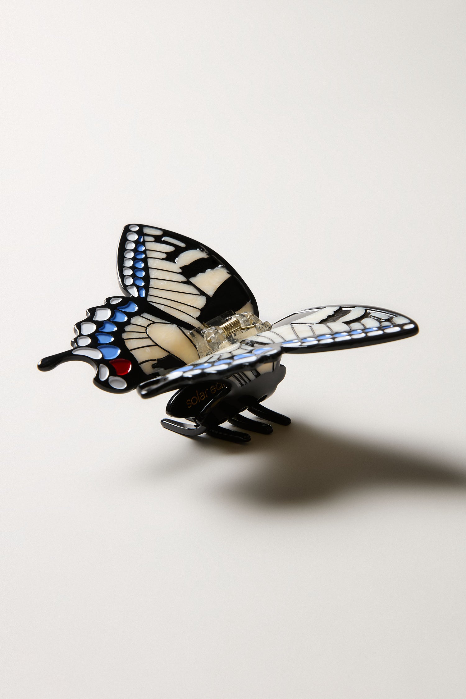 The Hand-Painted Swallowtail Hair Clip, featuring black, white, blue, and red accents with a gold-tone clip, rests on a light surface casting a gentle shadow.