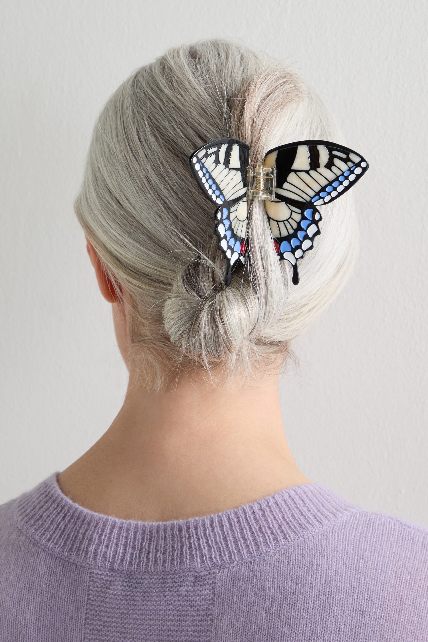 A person with gray hair in a low bun, secured with the Hand-Painted Swallowtail Hair Clip, is shown from behind against a plain background.