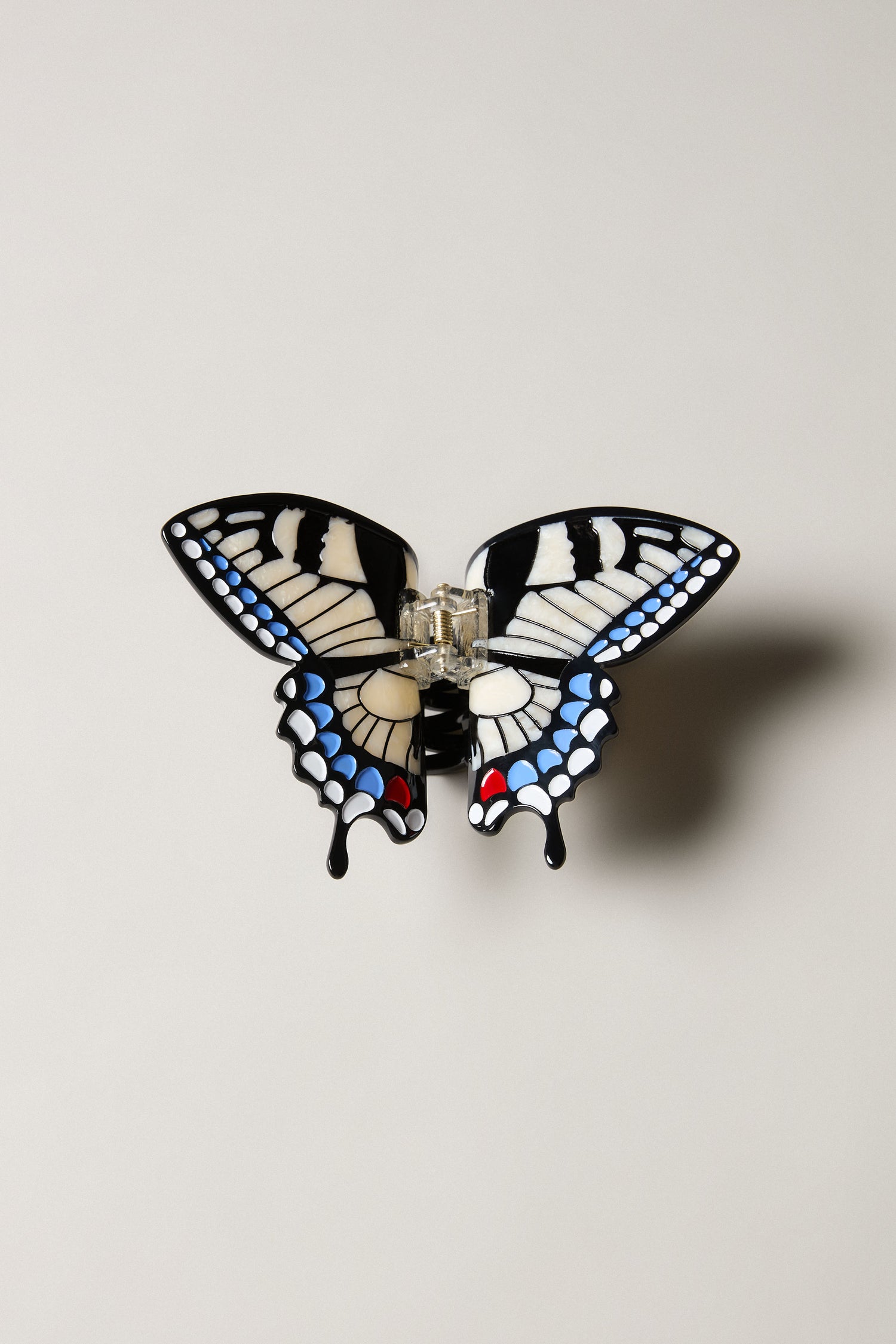 The Hand-Painted Swallowtail Hair Clip is a butterfly-shaped accessory with black, white, blue, and red patterns on a gold-tone clip, inspired by the swallowtail butterfly and shown against a light background.